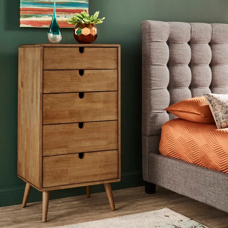 A mid-century modern wooden dresser stands next to a grey tufted bed with orange bedding against a deep green wall.