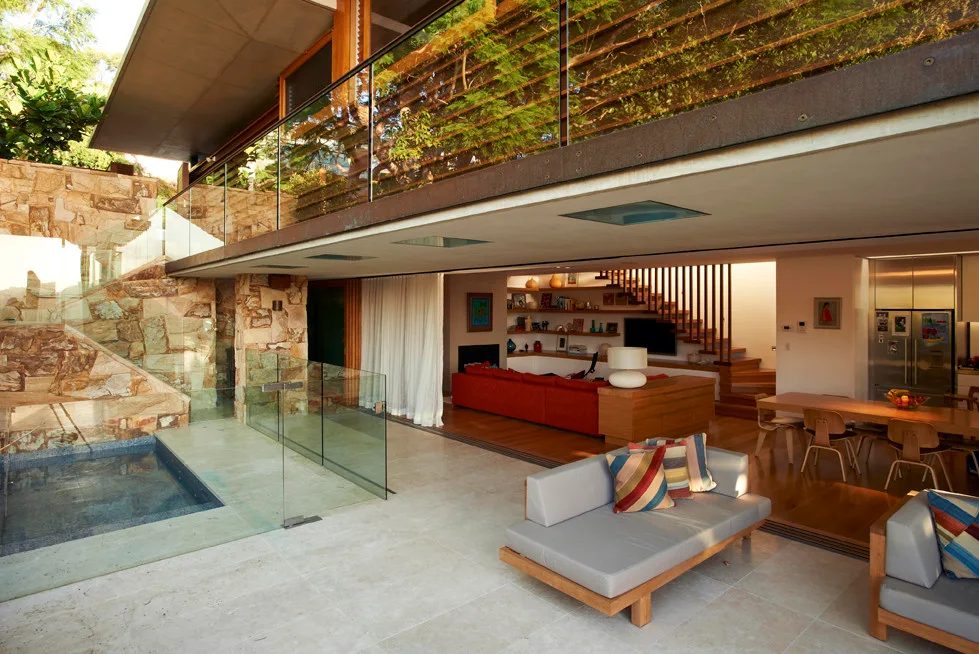 A modern open-plan home features a stone patio with a small pool that connects seamlessly to an indoor living area with a red sectional.