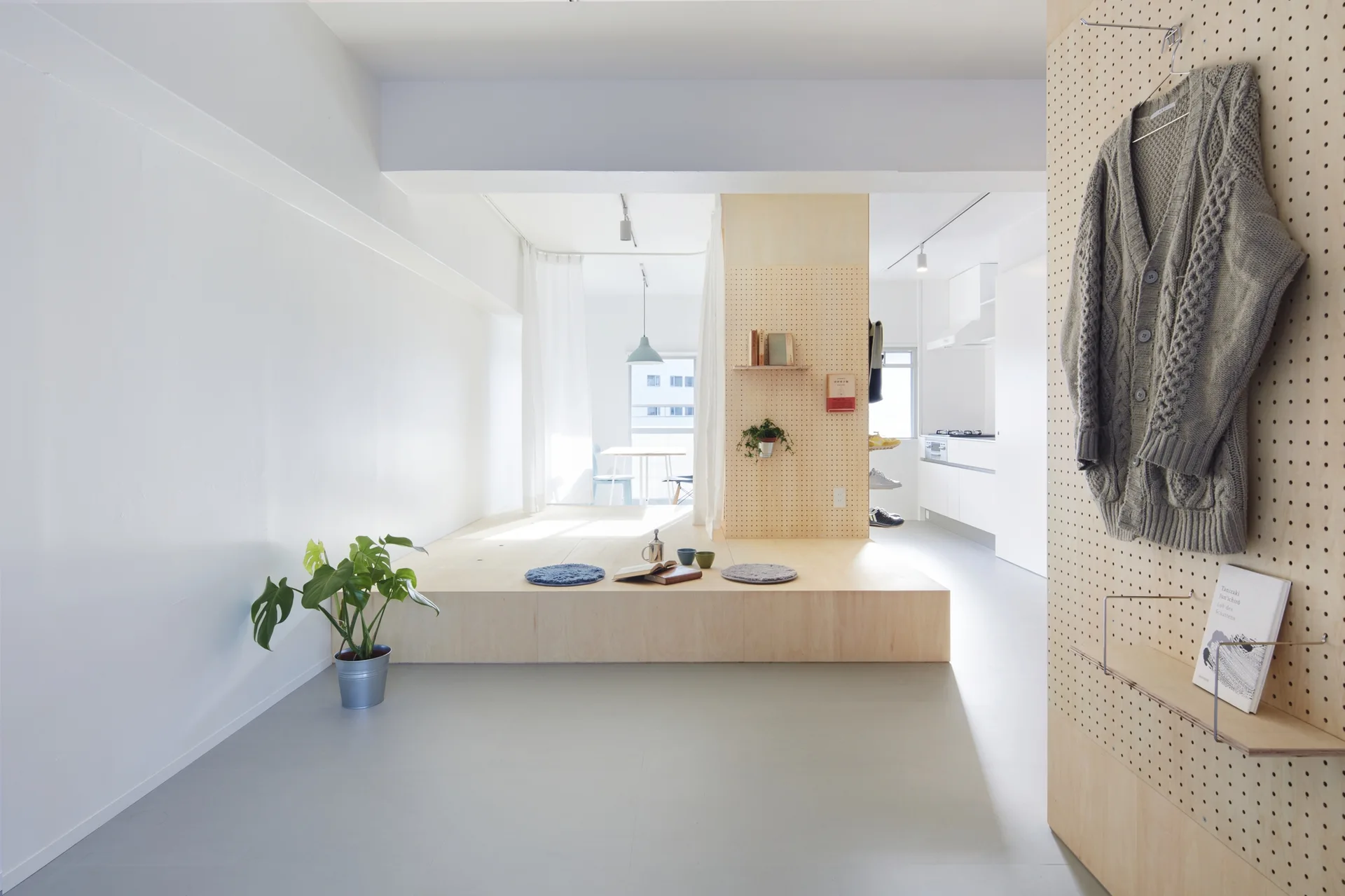 A minimalist apartment interior featuring a raised plywood platform, white walls, and a large wooden pegboard wall used for hanging clothes and shelves.