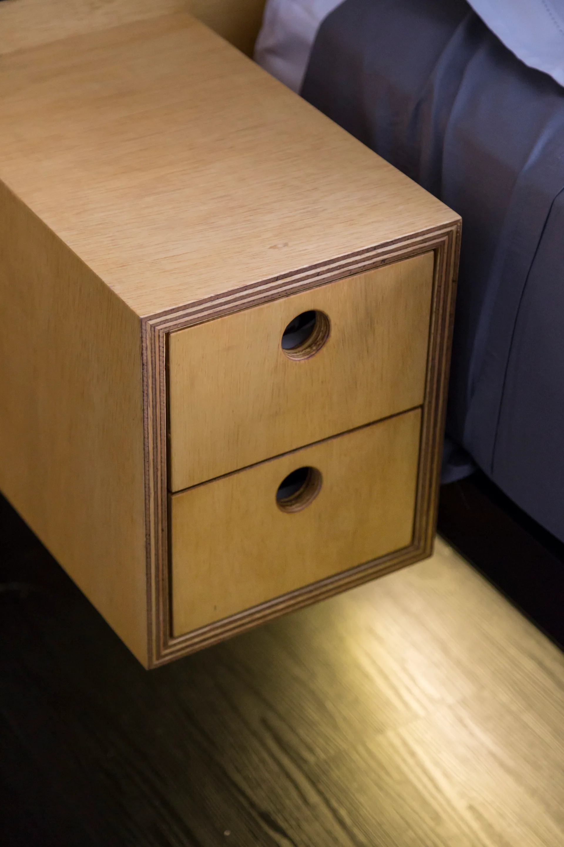 A wall-mounted plywood nightstand with two drawers featuring circular cutout pulls hangs beside a bed.