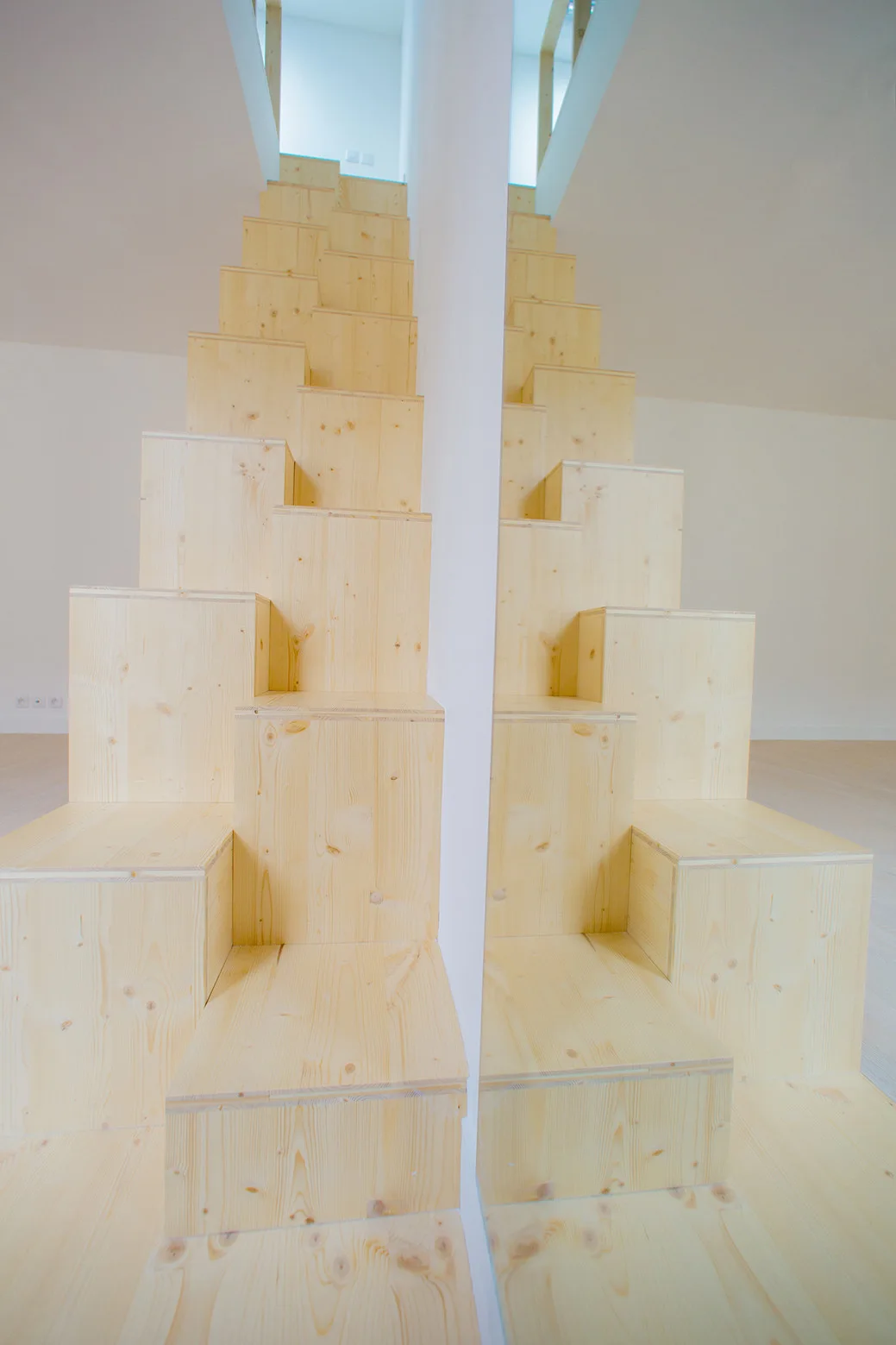 A modern alternating tread staircase made of stacked light wood blocks stands against a white wall next to a central mirror.