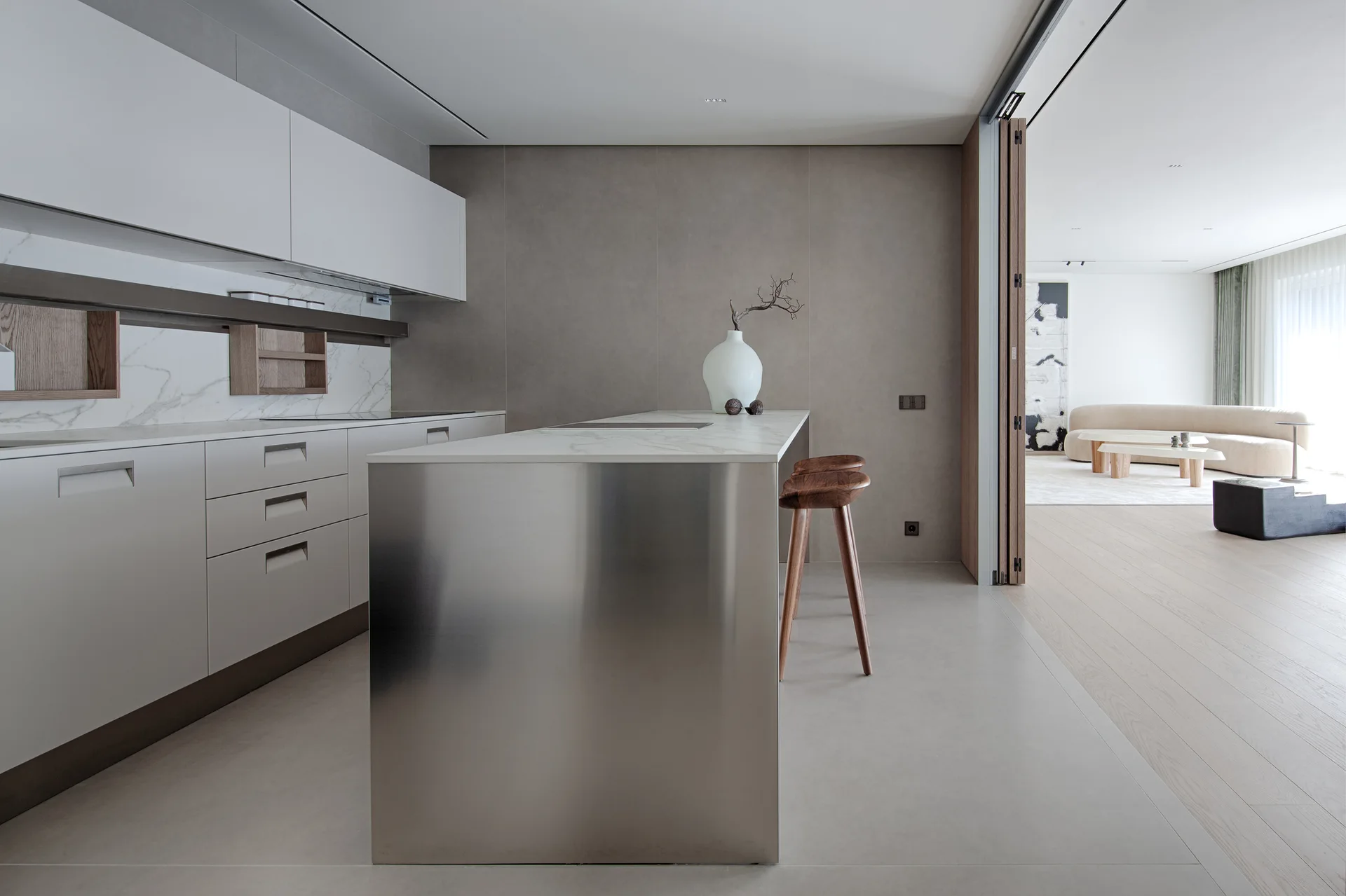 A minimalist kitchen features a stainless steel island with a marble top, white handleless cabinets, and a folding wooden door leading to a living room.