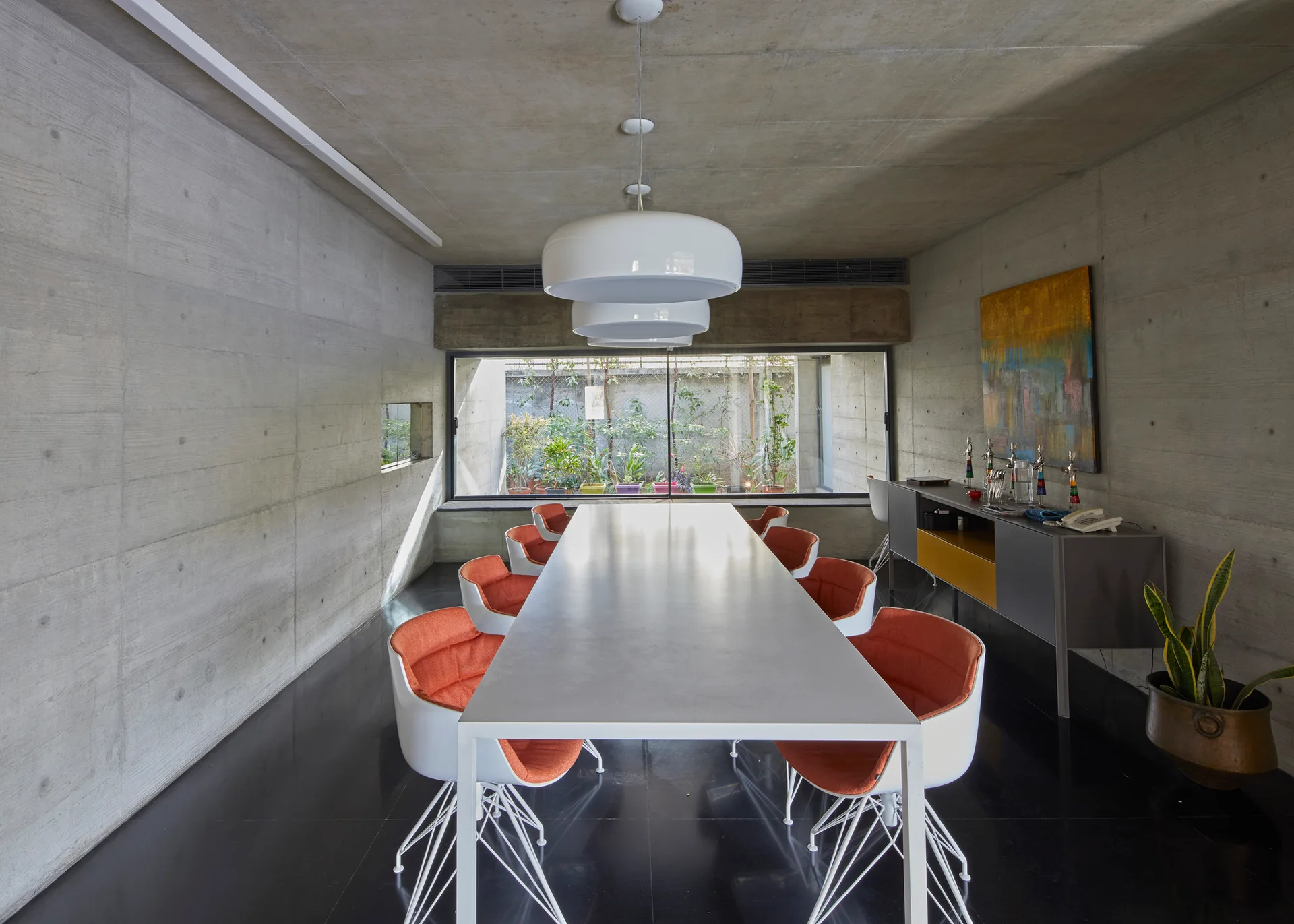 A long white dining table with orange chairs is set in a brutalist room with exposed concrete walls and a low window viewing a garden.