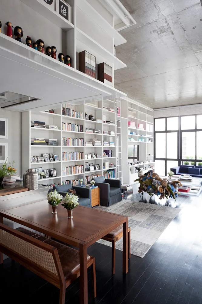 A modern industrial loft space with extensive white built-in bookshelves, a library ladder, a dark wood dining table, and an exposed concrete ceiling.