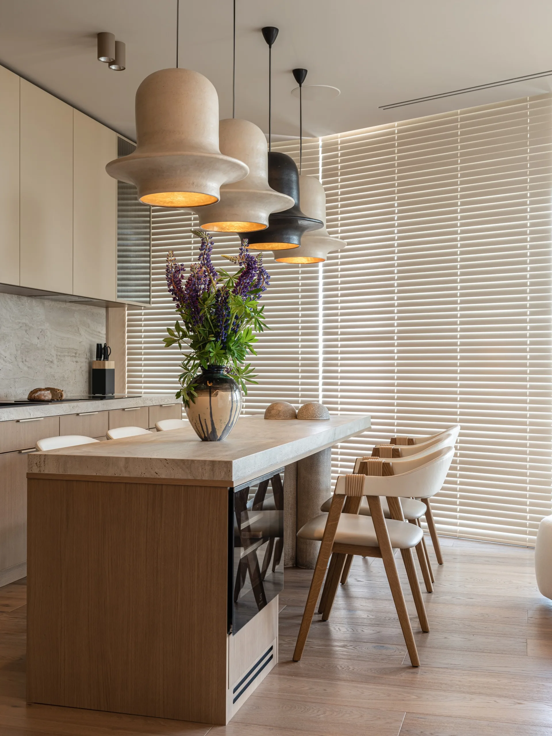 A modern kitchen island with a stone countertop features a vase of purple flowers, wooden counter stools, and a row of large sculptural pendant lights.