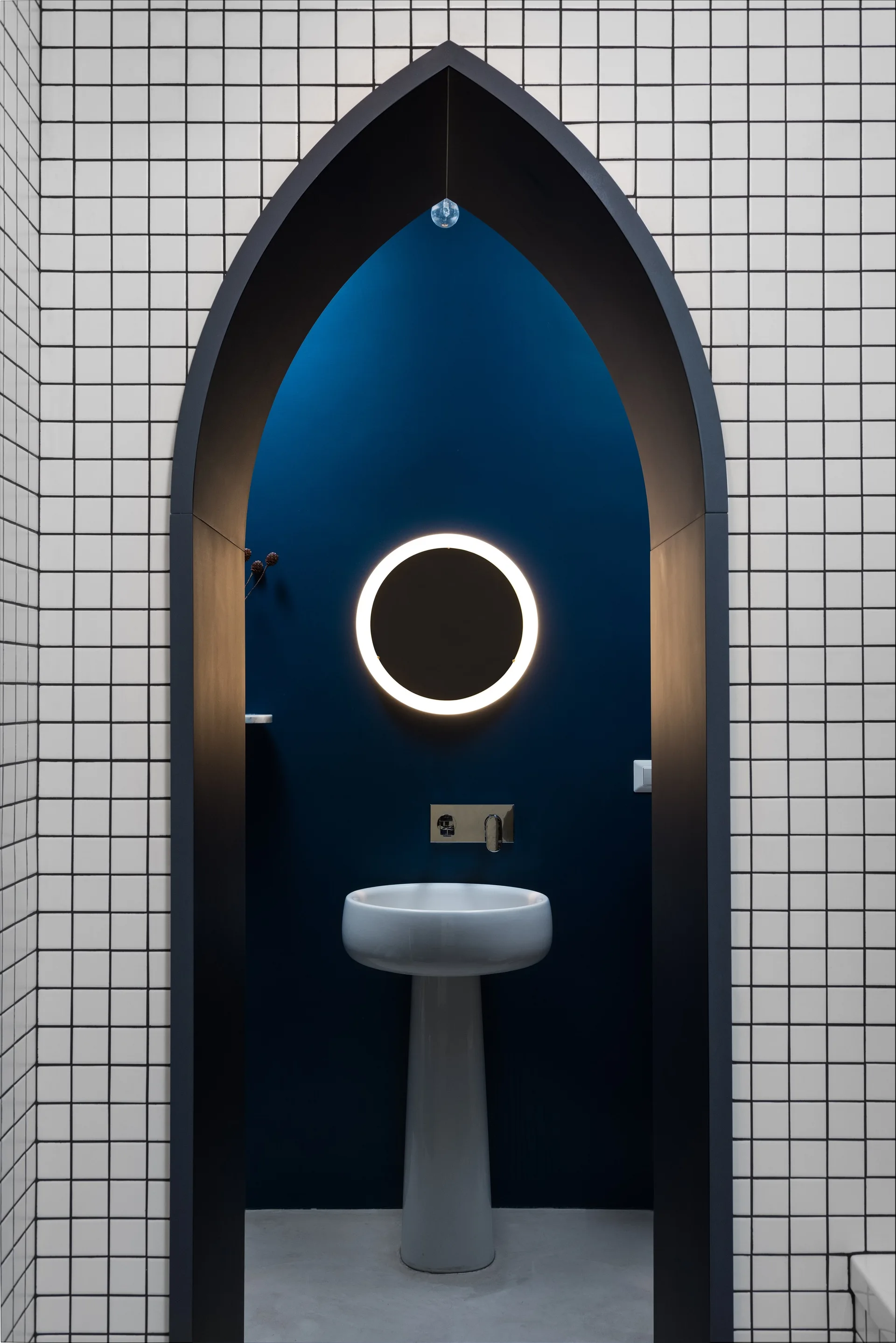 A white pedestal sink and backlit circular mirror set inside a deep blue pointed archway alcove surrounded by grid tiles.