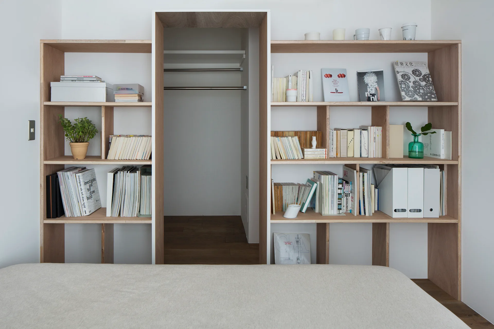 A light wood built-in shelving unit filled with books surrounds the open doorway of a walk-in closet in a minimalist bedroom.