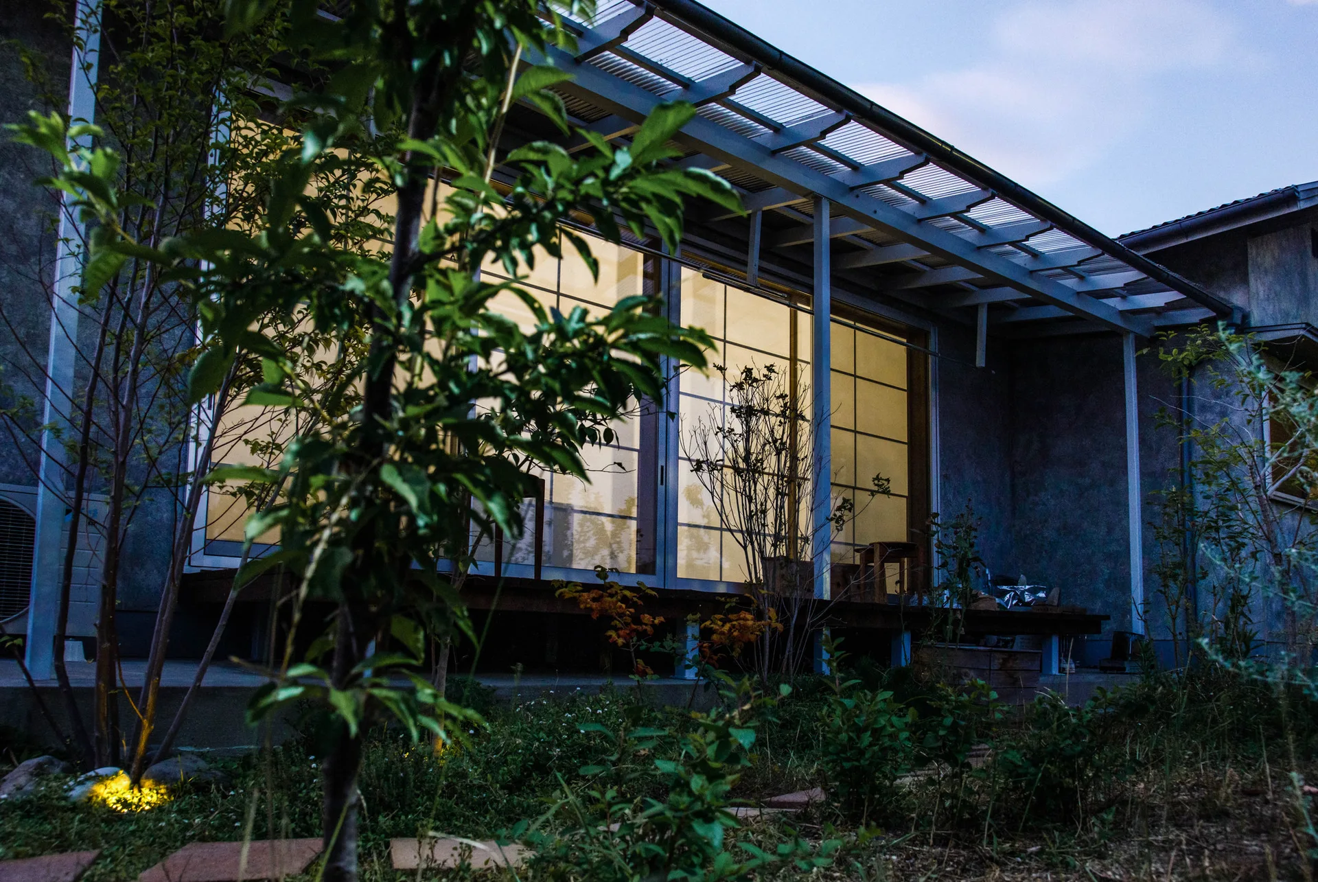 A wooden house deck features large sliding glass doors with a grid pattern glowing with warm light next to a natural garden at dusk.