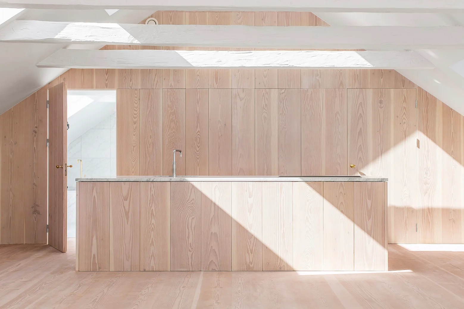 A minimalist attic kitchen features light wood paneled walls, a matching wood kitchen island, and white exposed ceiling beams.