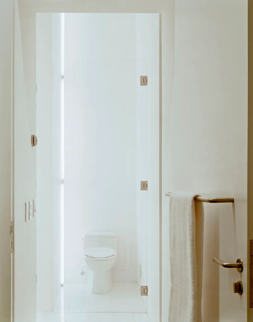 A view through an open doorway into a minimalist, all-white powder room featuring a simple ceramic toilet and metallic door hardware.