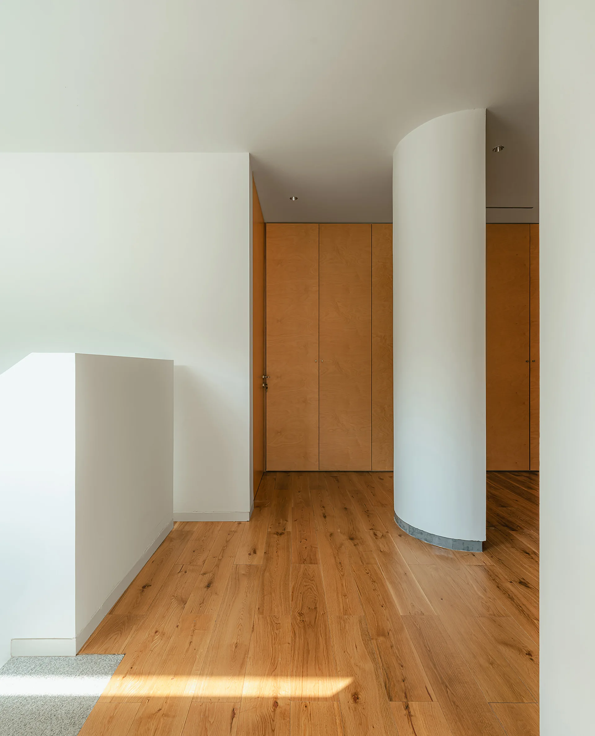 A minimalist hallway features light wood plank flooring, a curved white partition wall, and floor-to-ceiling wood paneled closet doors.
