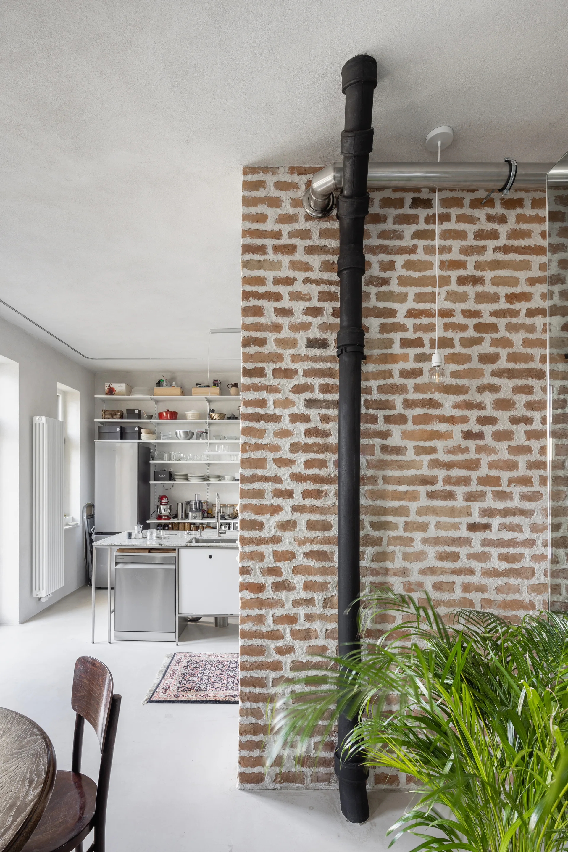 An industrial-style kitchen seen past an exposed red brick partition wall featuring a prominent vertical black pipe and open white shelving.