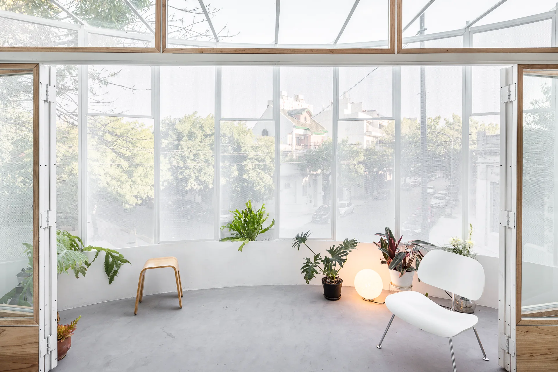 A minimalist sunroom features a curved wall of mesh-screened windows, a white modern lounge chair, a wooden stool, and several potted plants.