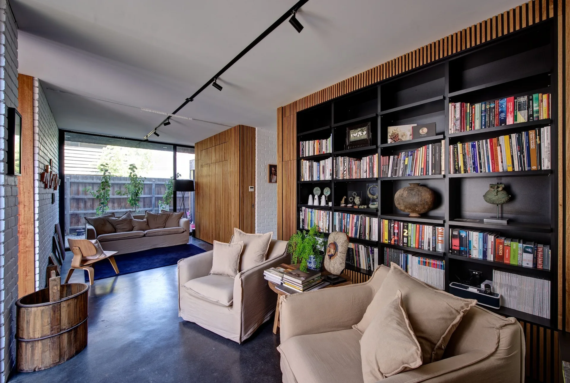 A modern living room features a tall black bookcase framed by vertical wood slats, beige armchairs, and a polished concrete floor.