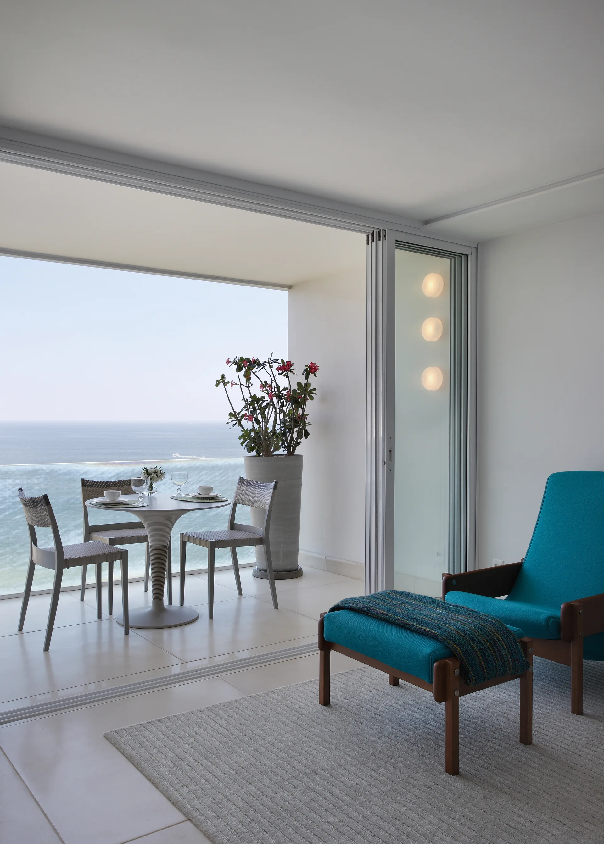 A modern apartment interior featuring a teal armchair with matching ottoman opens to a tiled balcony with a dining table and ocean views.