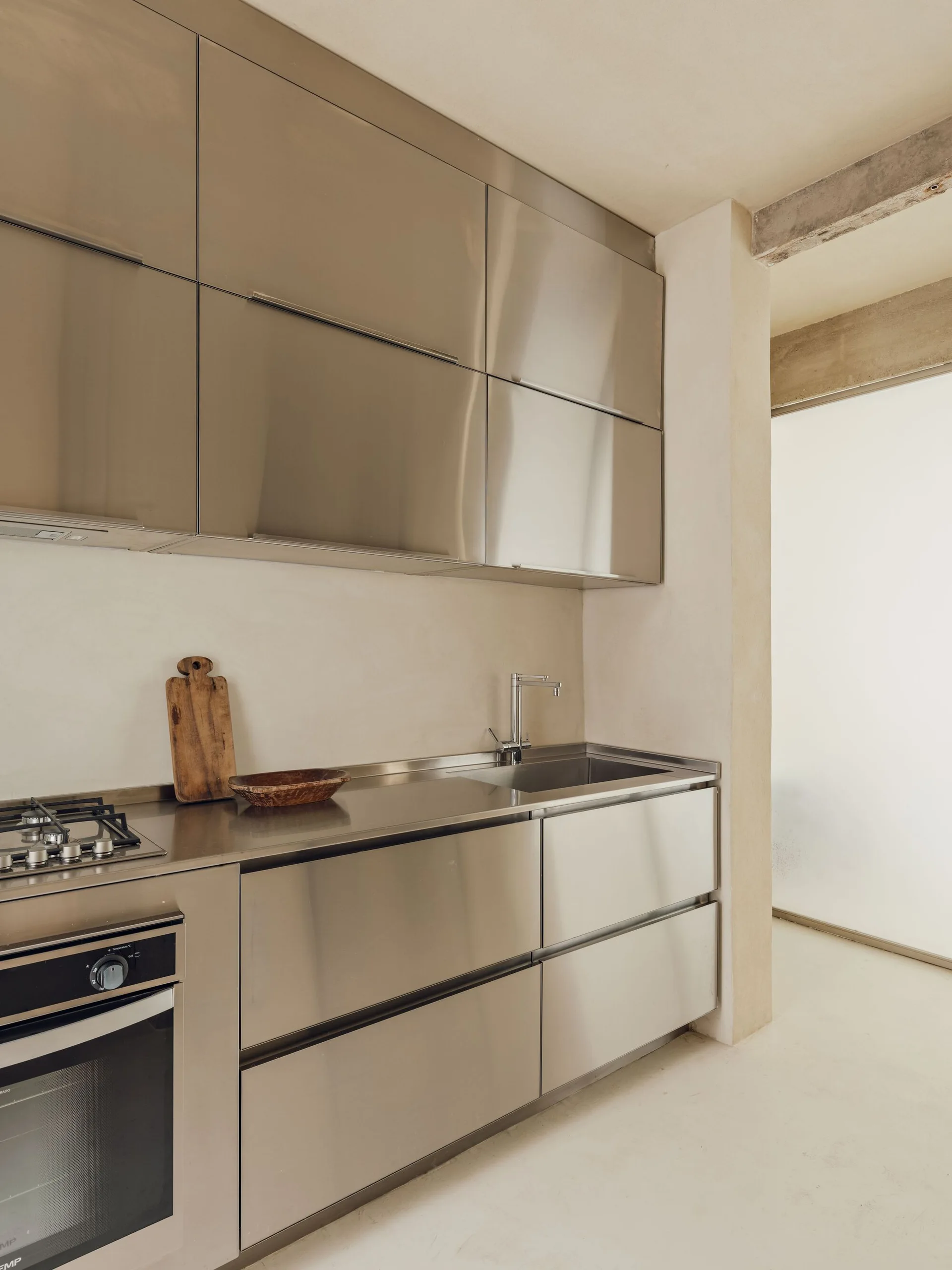A minimalist kitchen featuring stainless steel upper and lower cabinets, an integrated metal countertop with a sink, and textured beige plaster walls.