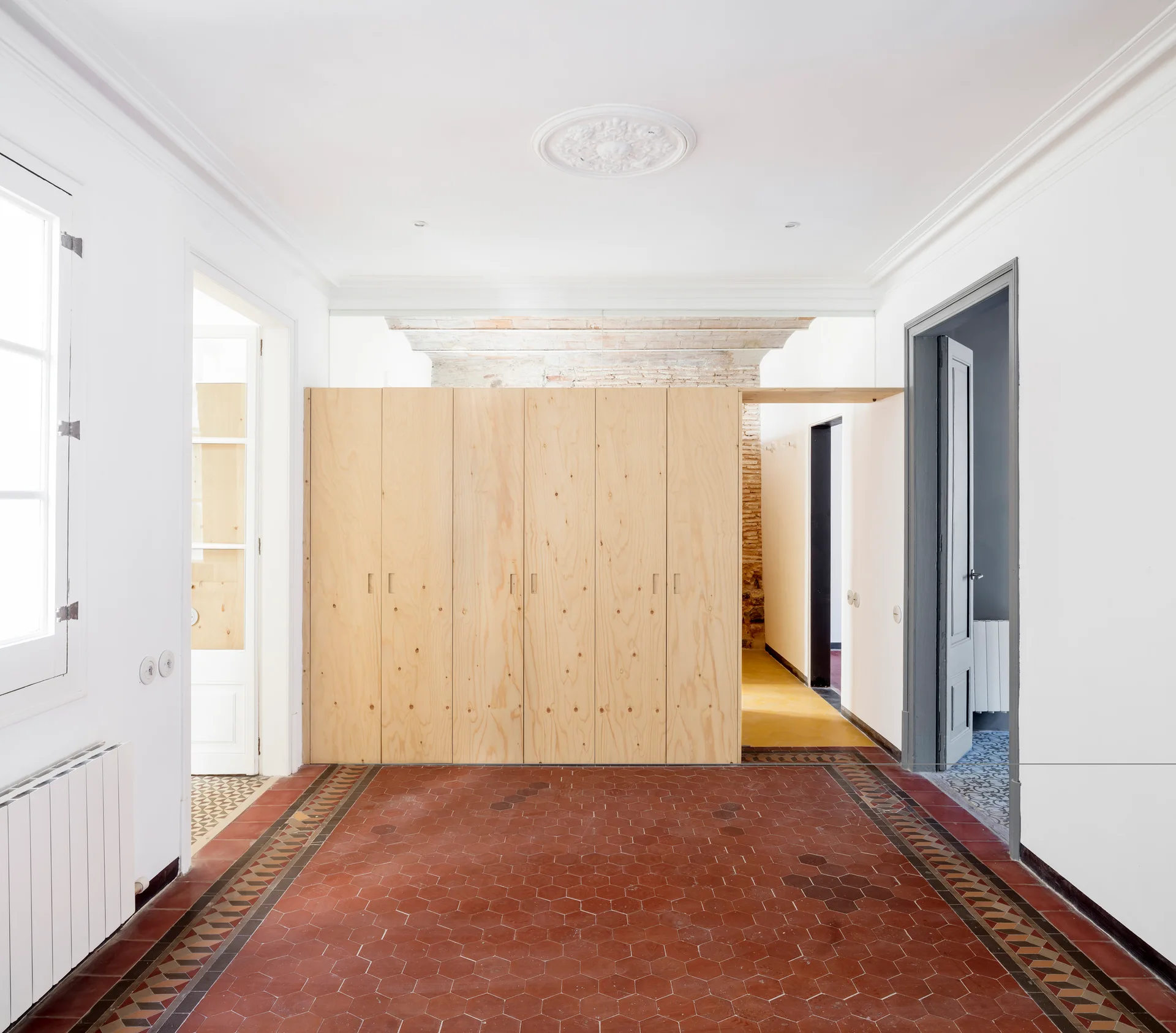 A bright hallway showcases red hexagonal tile floors, white walls, and a large built-in light wood storage cabinet.