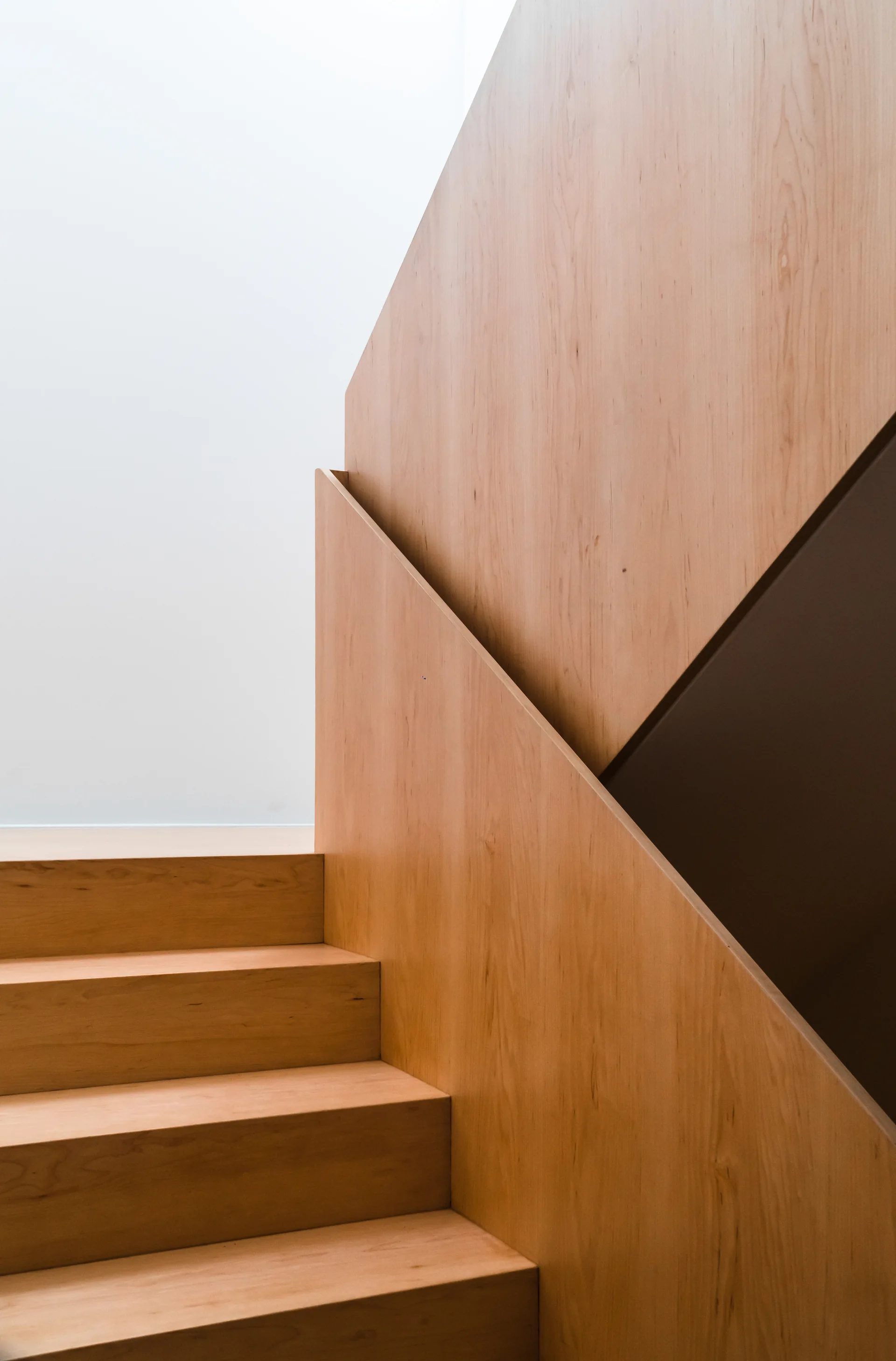 A minimalist wooden staircase features light-colored steps, matching wood wall paneling, and an integrated geometric handrail against a plain white wall.