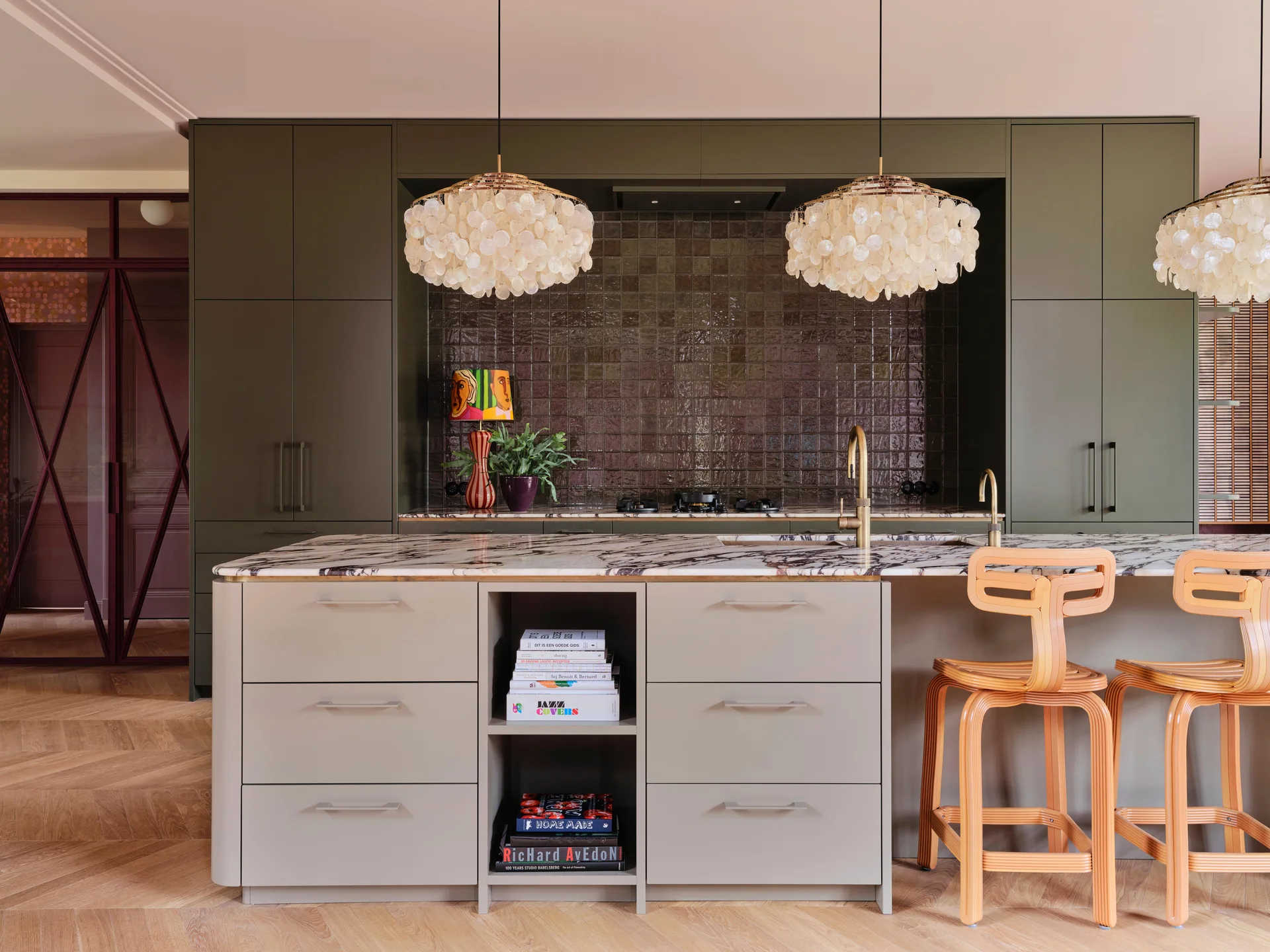 A modern kitchen with matte olive green cabinetry, a glossy dark tile backsplash, and a marble island with wooden stools under shell pendant lights.