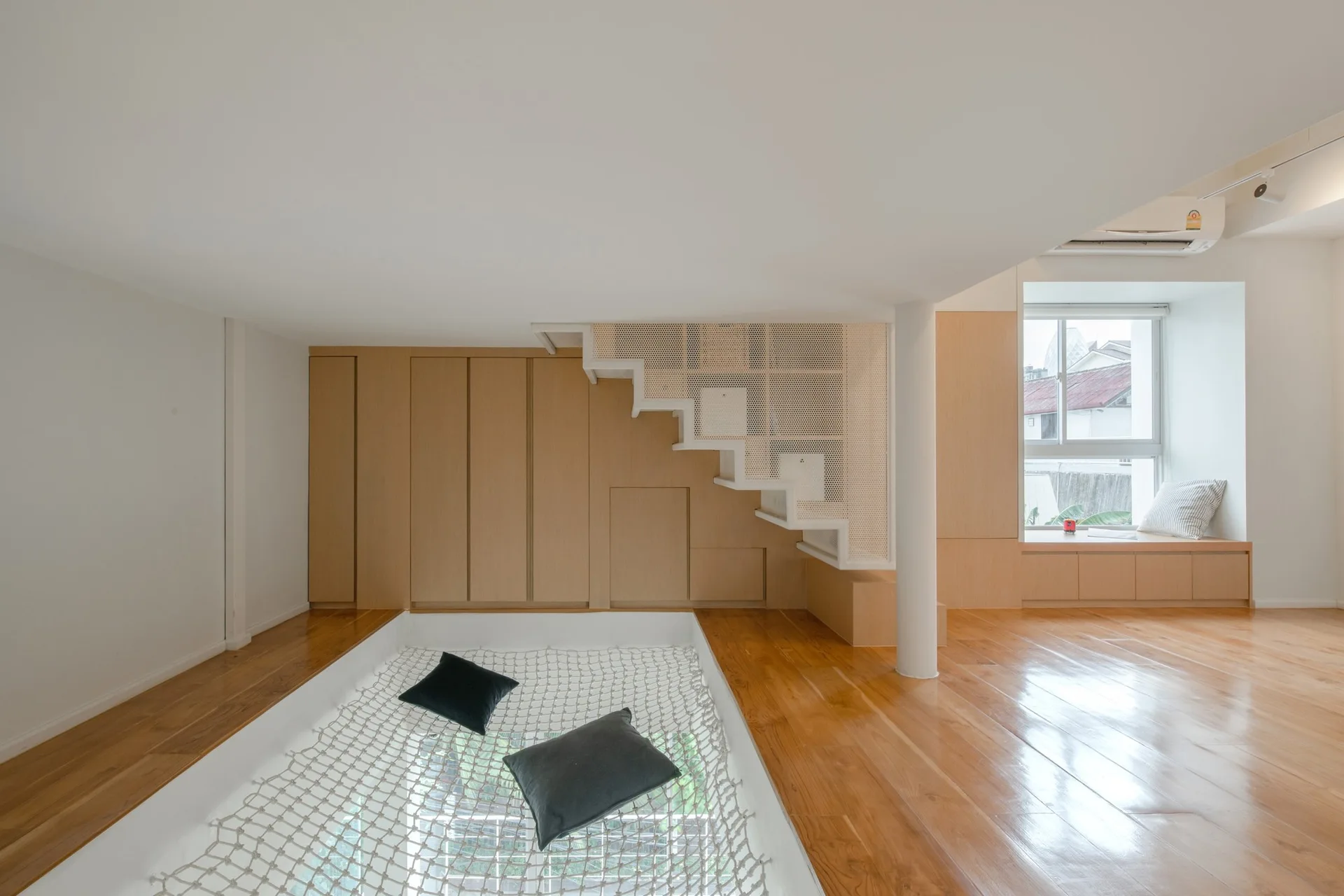 A minimalist loft space features a white metal mesh staircase, light wood cabinetry, and a sunken net lounge with two dark grey pillows.