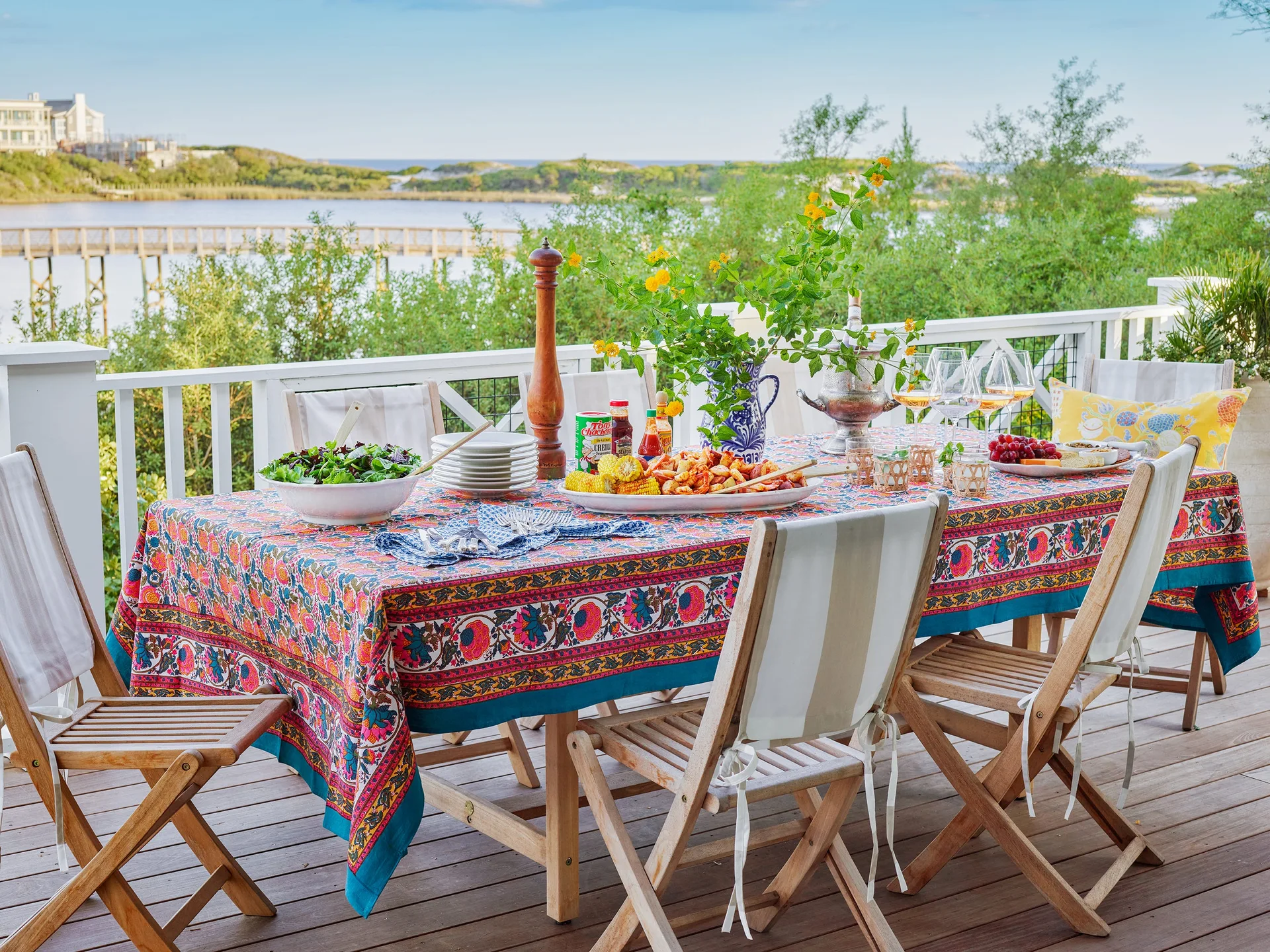 An outdoor wooden dining table set with a colorful floral tablecloth and folding chairs sits on a deck overlooking a coastal landscape.
