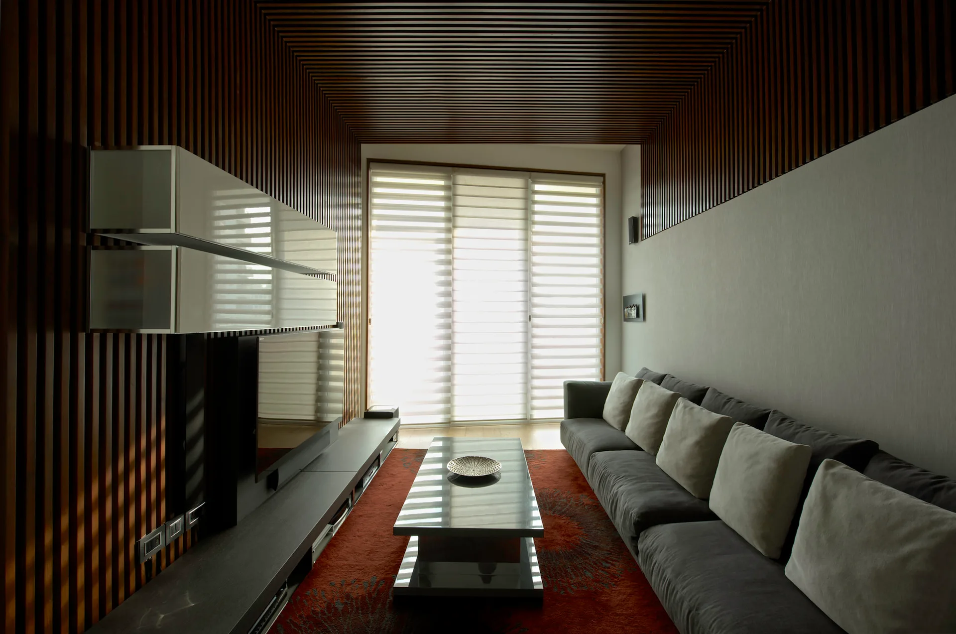 A narrow modern living room with dark wood slat walls and ceiling, a long grey sofa, and a large window with horizontal blinds.