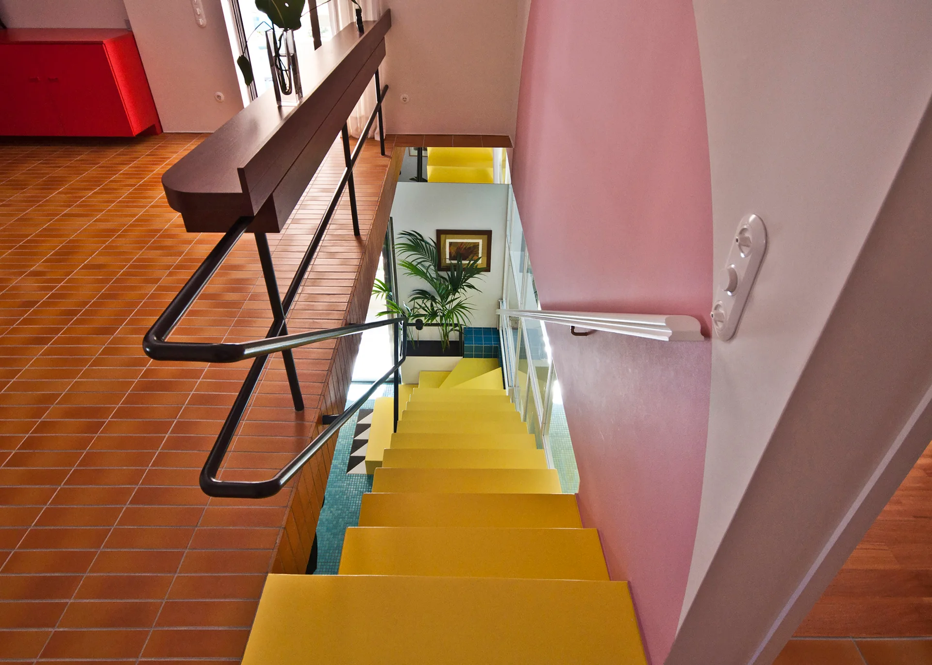 A view down a bright yellow staircase with a pink color-blocked accent wall and terracotta tile flooring.