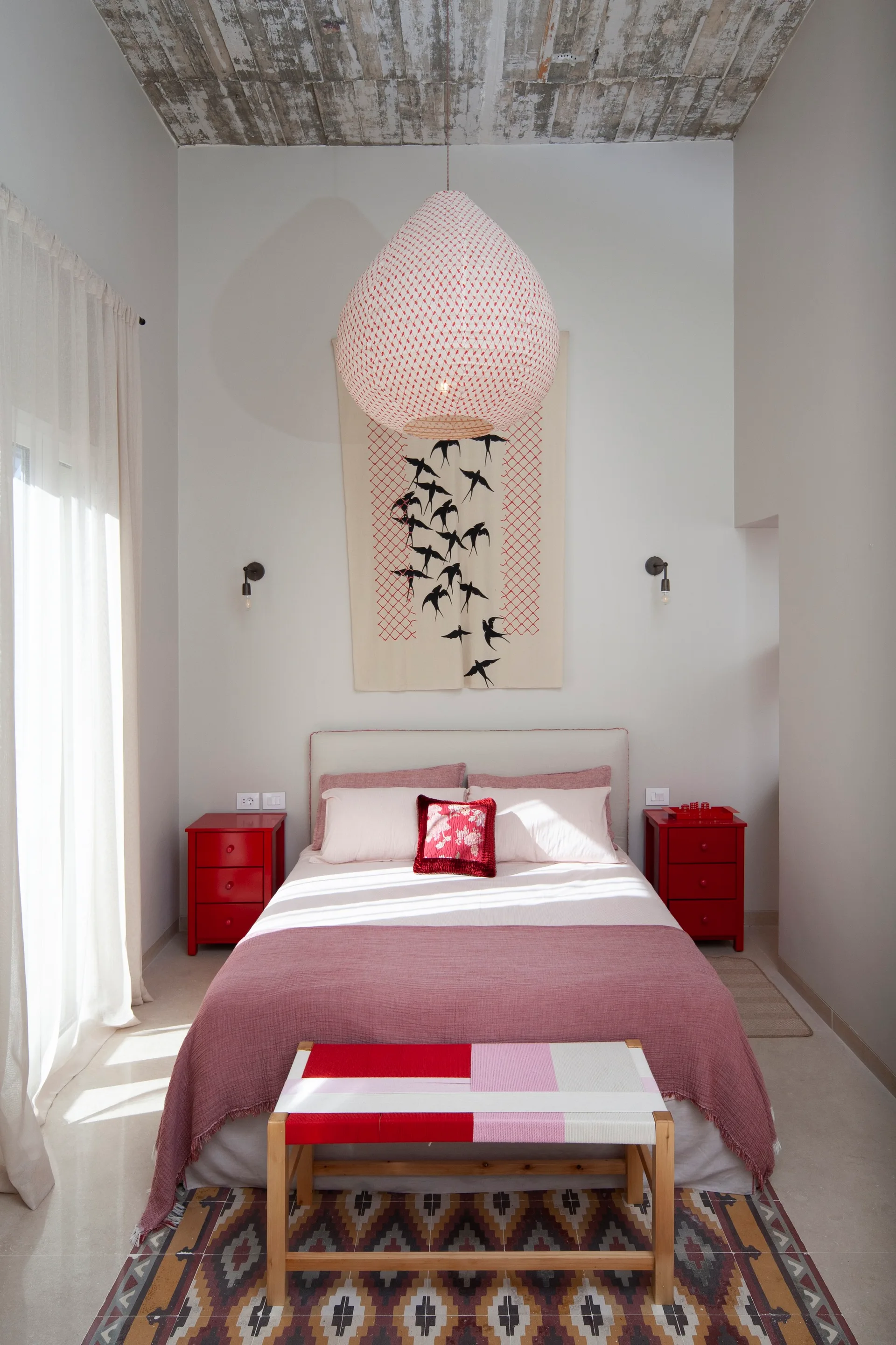 A modern bedroom features a bed flanked by glossy red nightstands beneath a large paper lantern light and a bird tapestry.