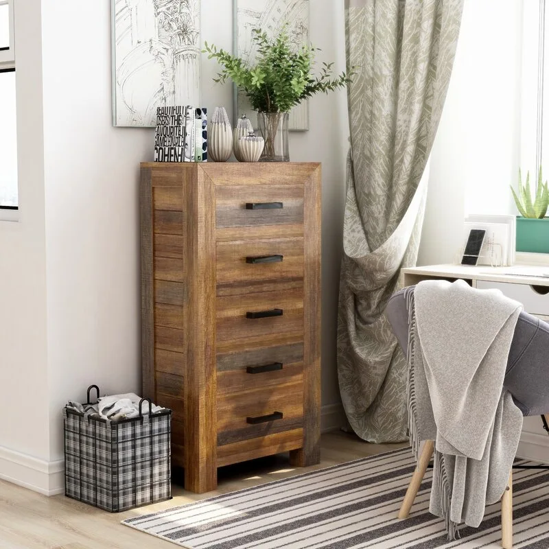 A tall rustic wood five-drawer chest stands on a striped rug next to a window draped with patterned curtains and a small desk with a chair.