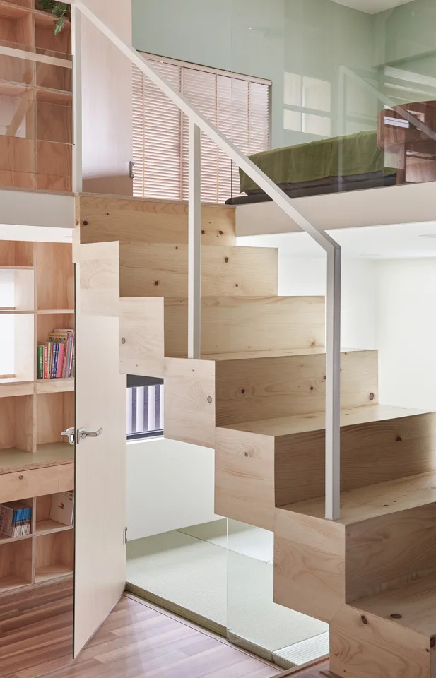 A light wood block staircase with a white metal railing leads to a loft-level bedroom next to built-in plywood shelving.