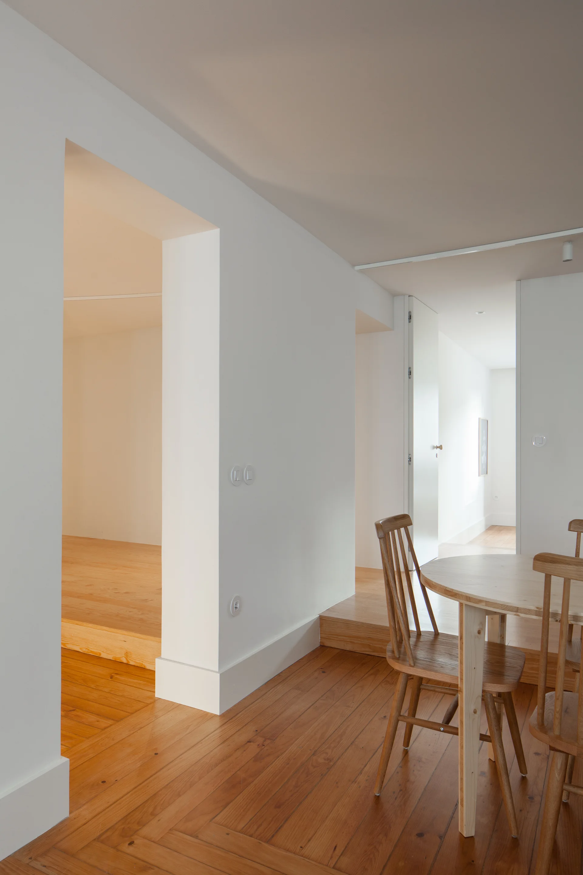 A minimalist dining area features a light wood circular table and spindle-back chairs set against clean white walls and warm wood floors.
