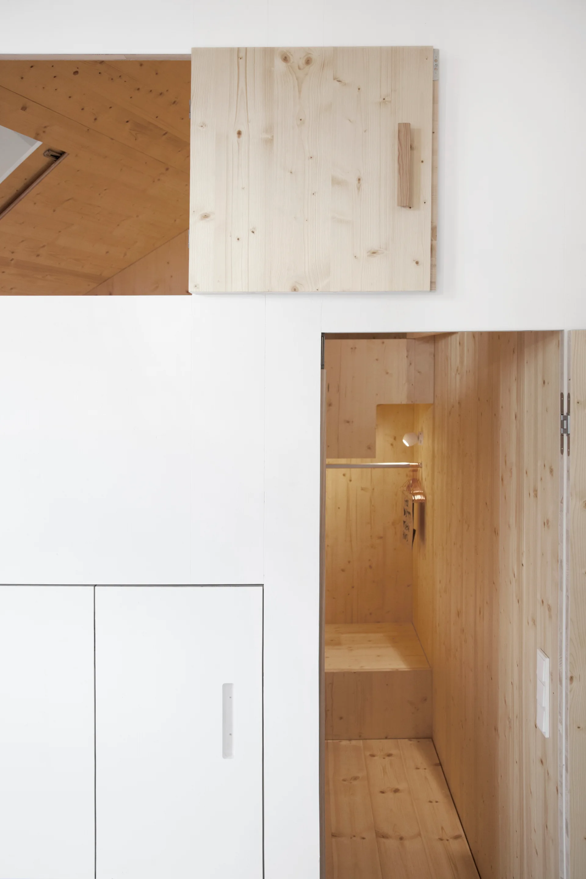 A minimalist white built-in storage wall featuring a recessed, natural wood-lined closet alcove with a metal hanging rod and a warm spotlight.