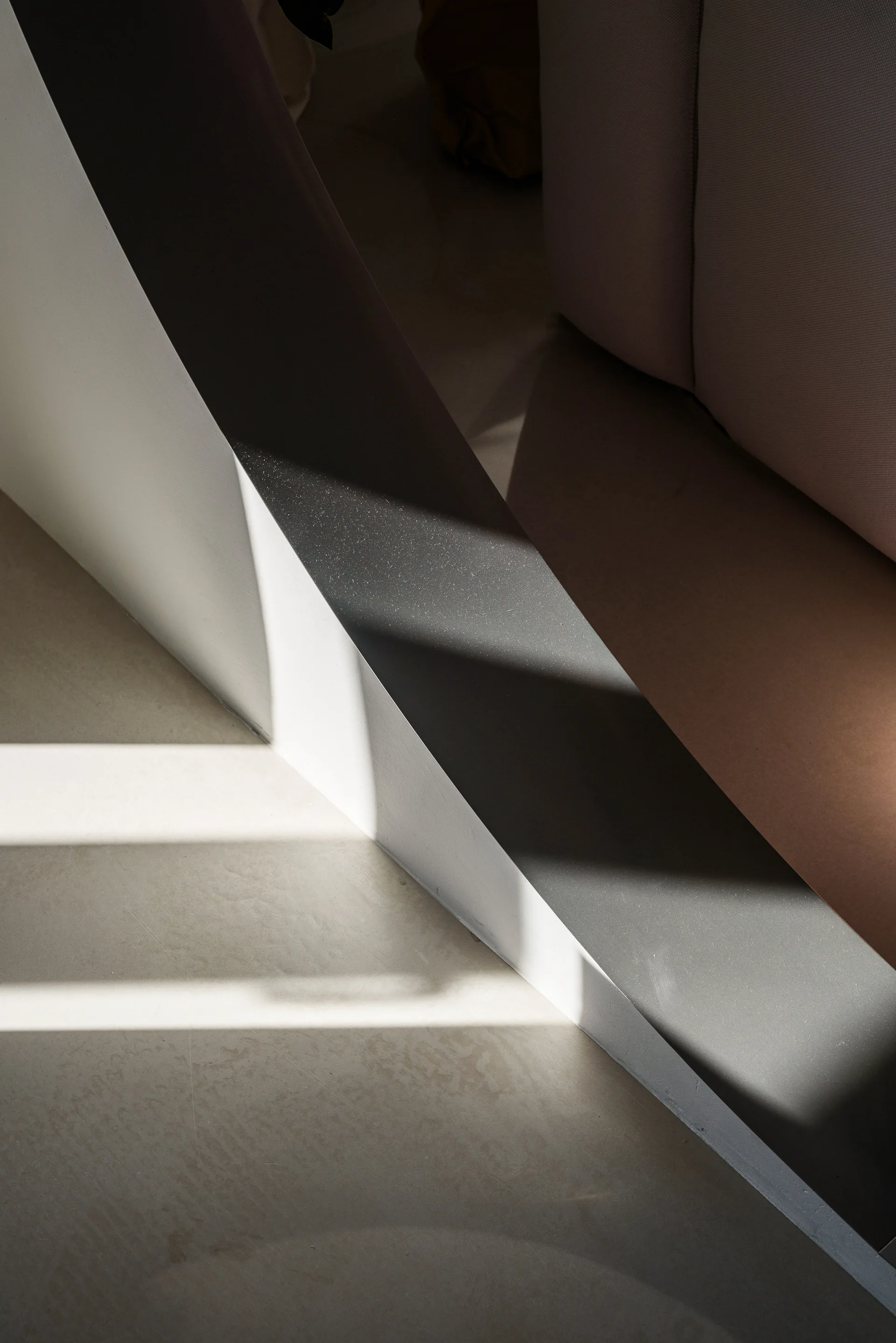 Geometric architectural lines cast sharp linear shadows across a smooth concrete floor next to a neutral-toned upholstered sofa corner.