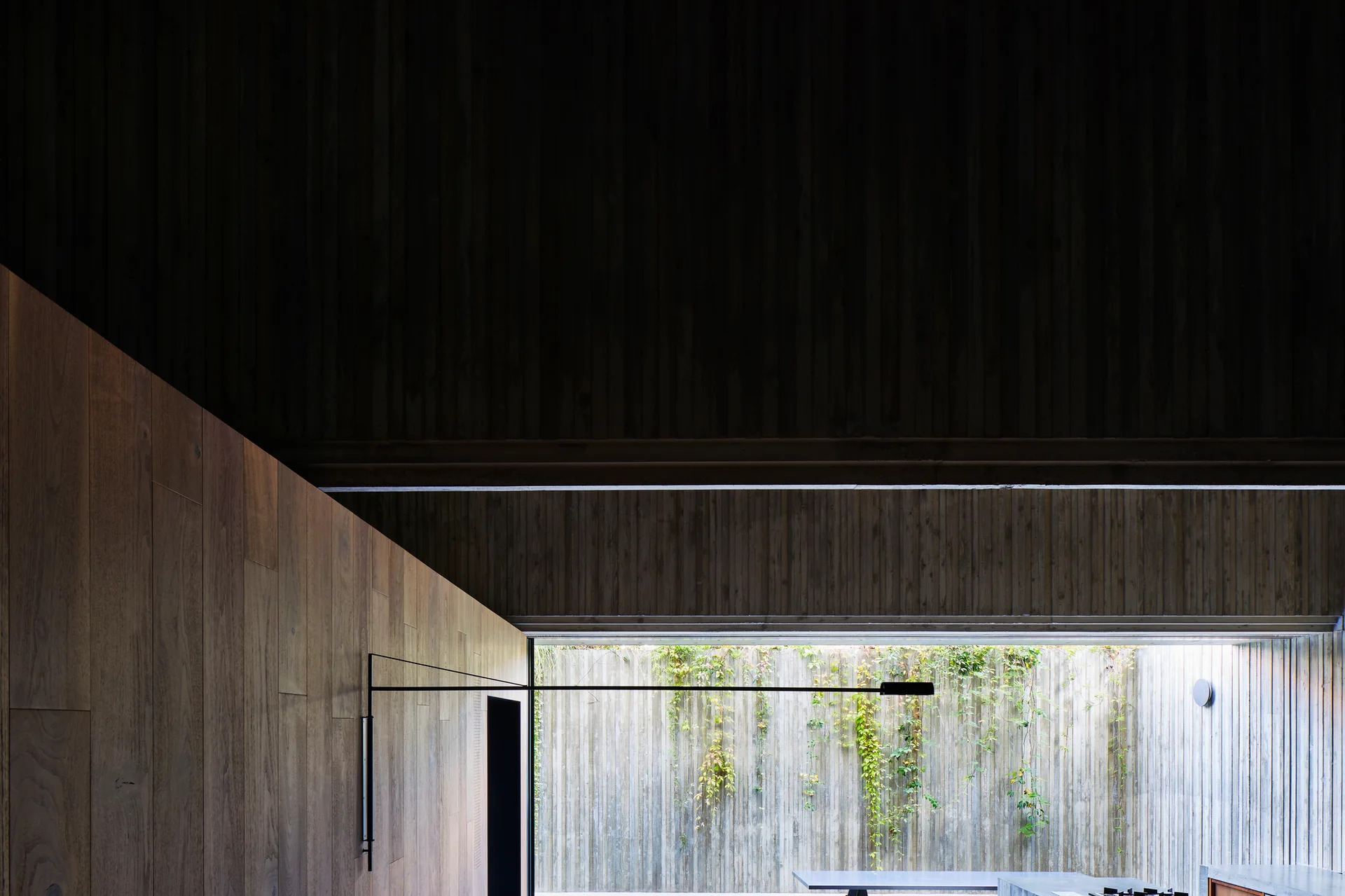 Minimalist room with wood paneled walls and a slim black sconce looking out to a concrete courtyard with climbing green vines.