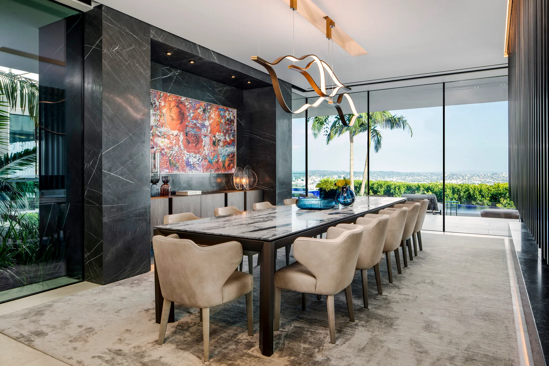 A modern dining room features a long marble table surrounded by beige chairs, set against a dark veined marble wall and large glass windows.