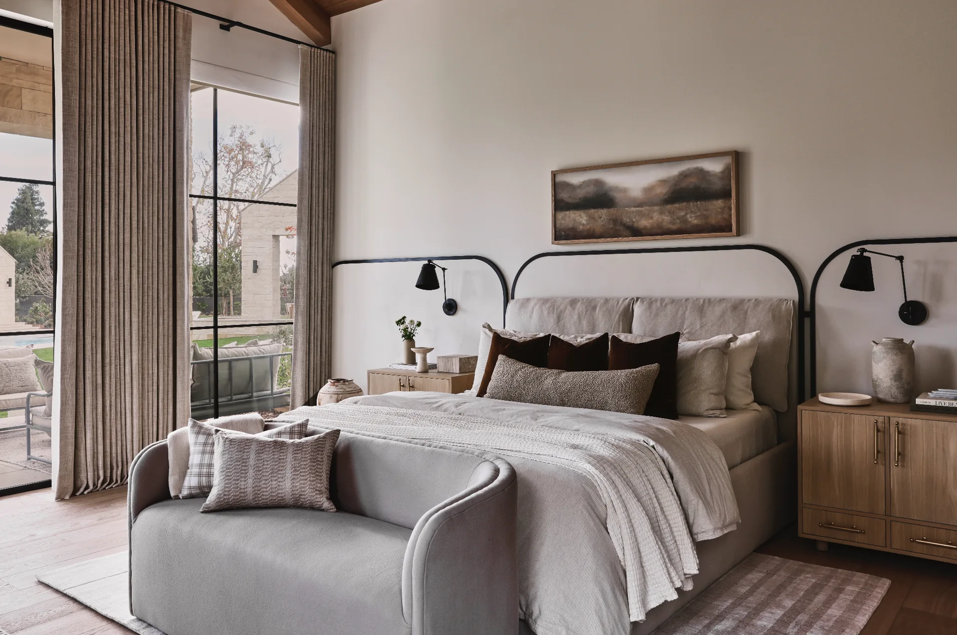 An upholstered bed with textured pillows sits between light wood nightstands, highlighted by a black metal wall frame and adjacent to large windows.