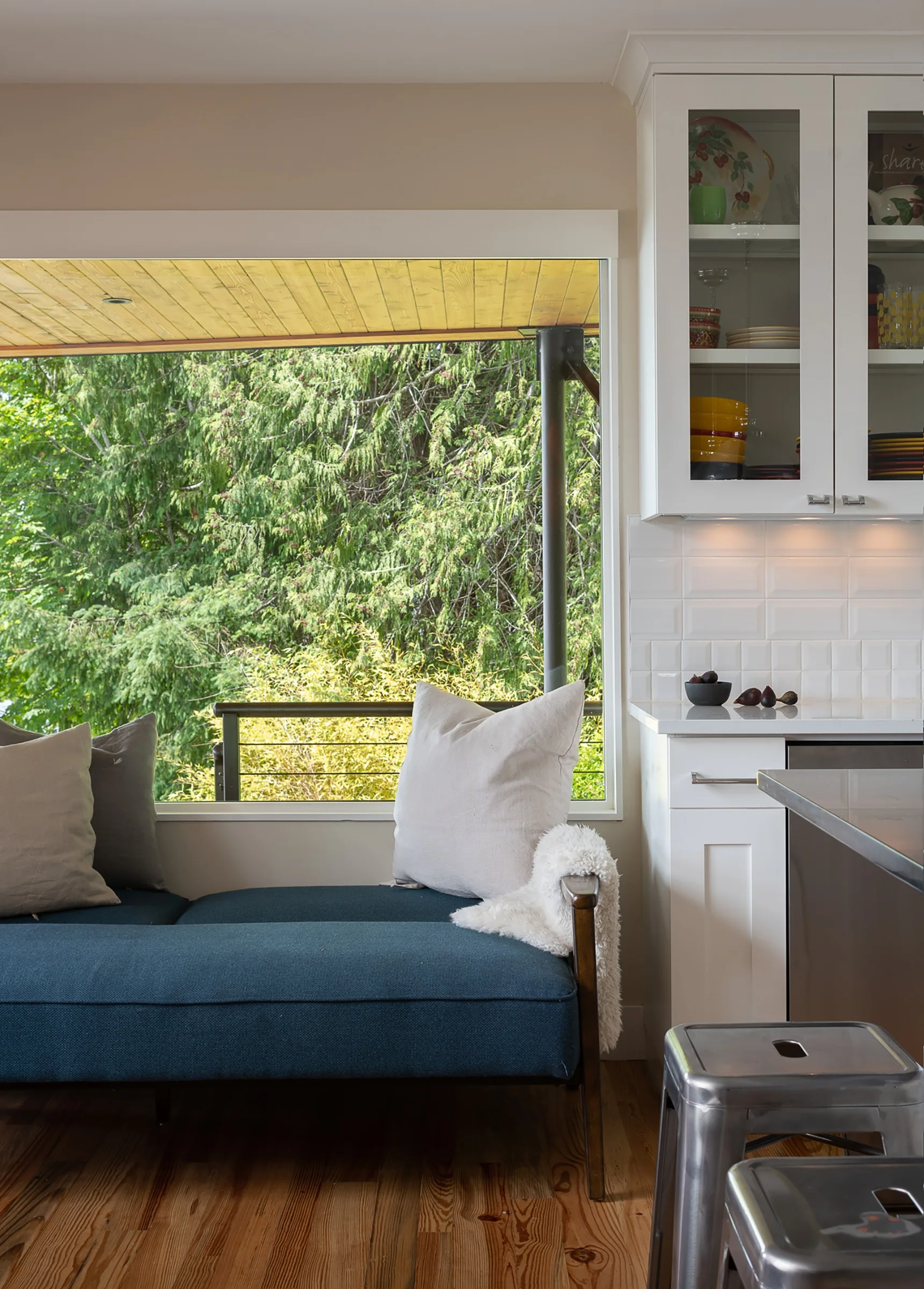A blue upholstered daybed sits beneath a large window next to white kitchen cabinets with glass doors and a subway tile backsplash.
