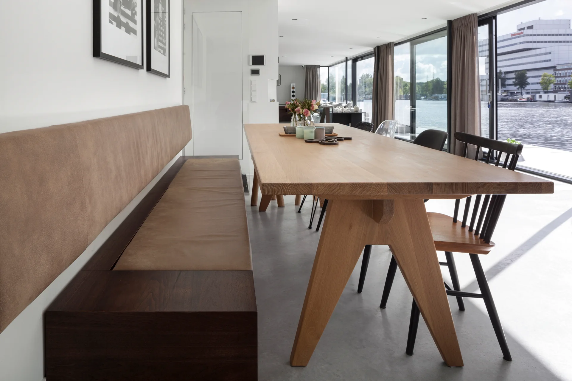 A light wood dining table paired with a built-in tan leather banquette and black spindle chairs in a room with floor-to-ceiling windows.