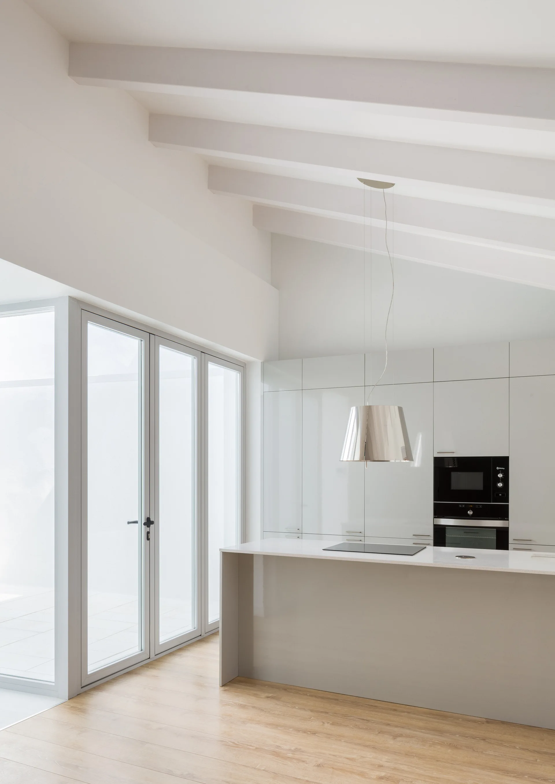 A minimalist white kitchen features a central island, high-gloss flat-panel cabinets, and a sloped ceiling with exposed beams.