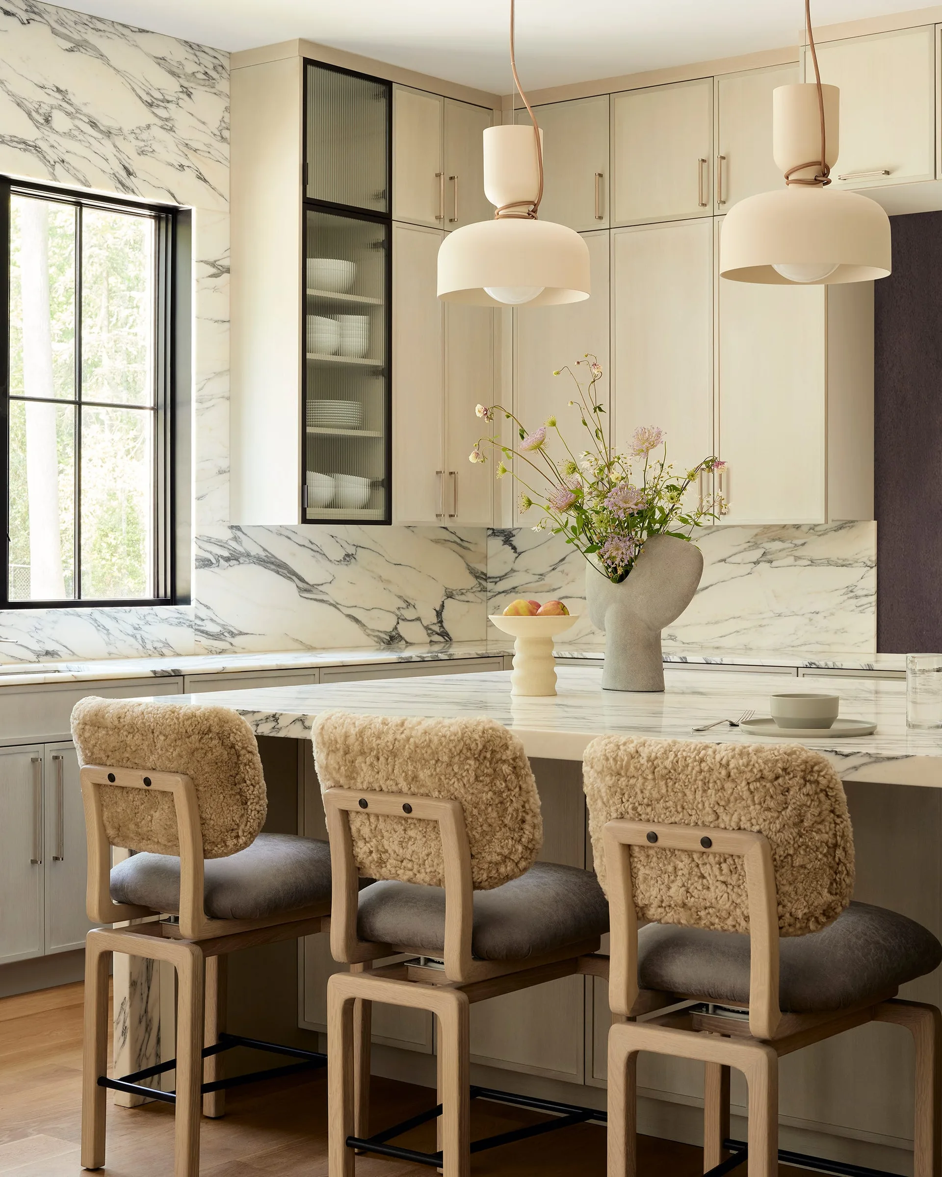 A modern kitchen features a marble-topped island with three shearling-backed wooden stools, beige cabinets, and a veined marble backsplash.