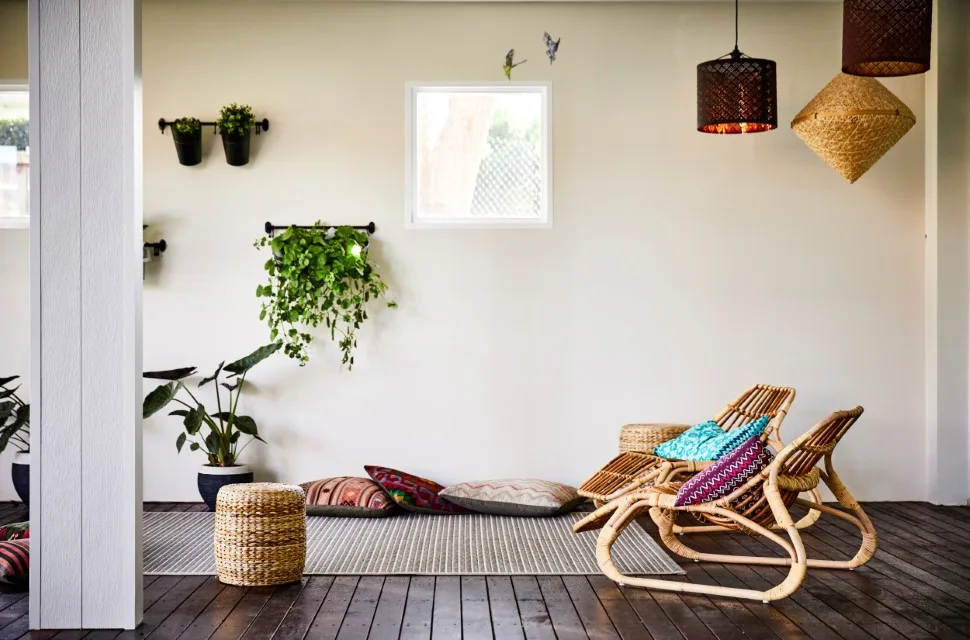 Curved rattan lounge chairs and patterned floor cushions sit on a dark wood deck next to wall-mounted planters and a square window.