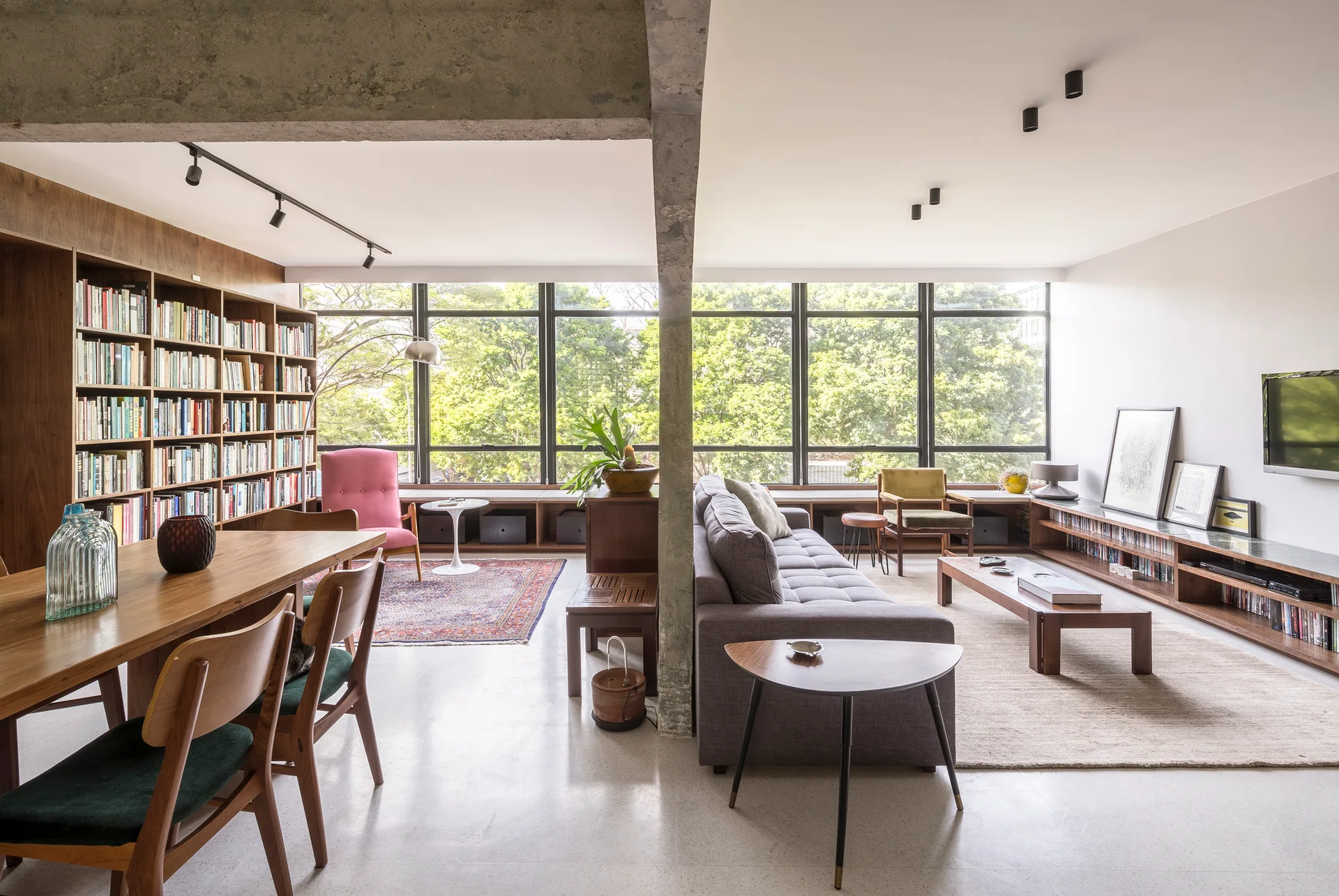 An open-concept living and dining room features a grey sofa, wooden table, large bookshelf, and a concrete column next to a wall of windows.