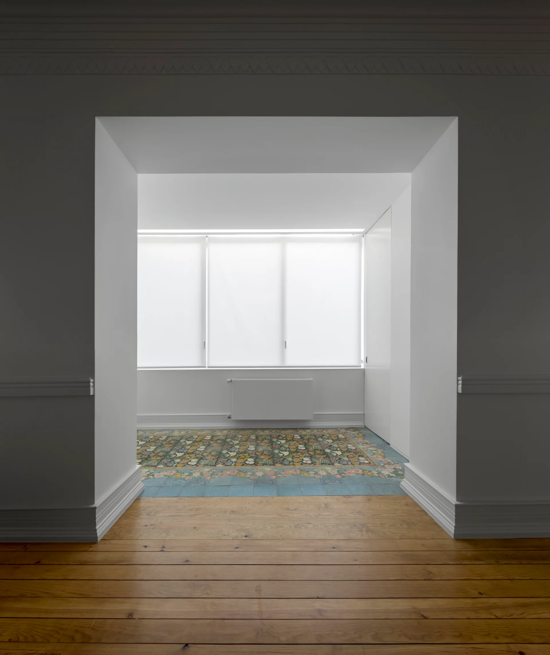 A view through a deep white cased opening from a natural wood floor into an empty room with colorful floral floor tiles.