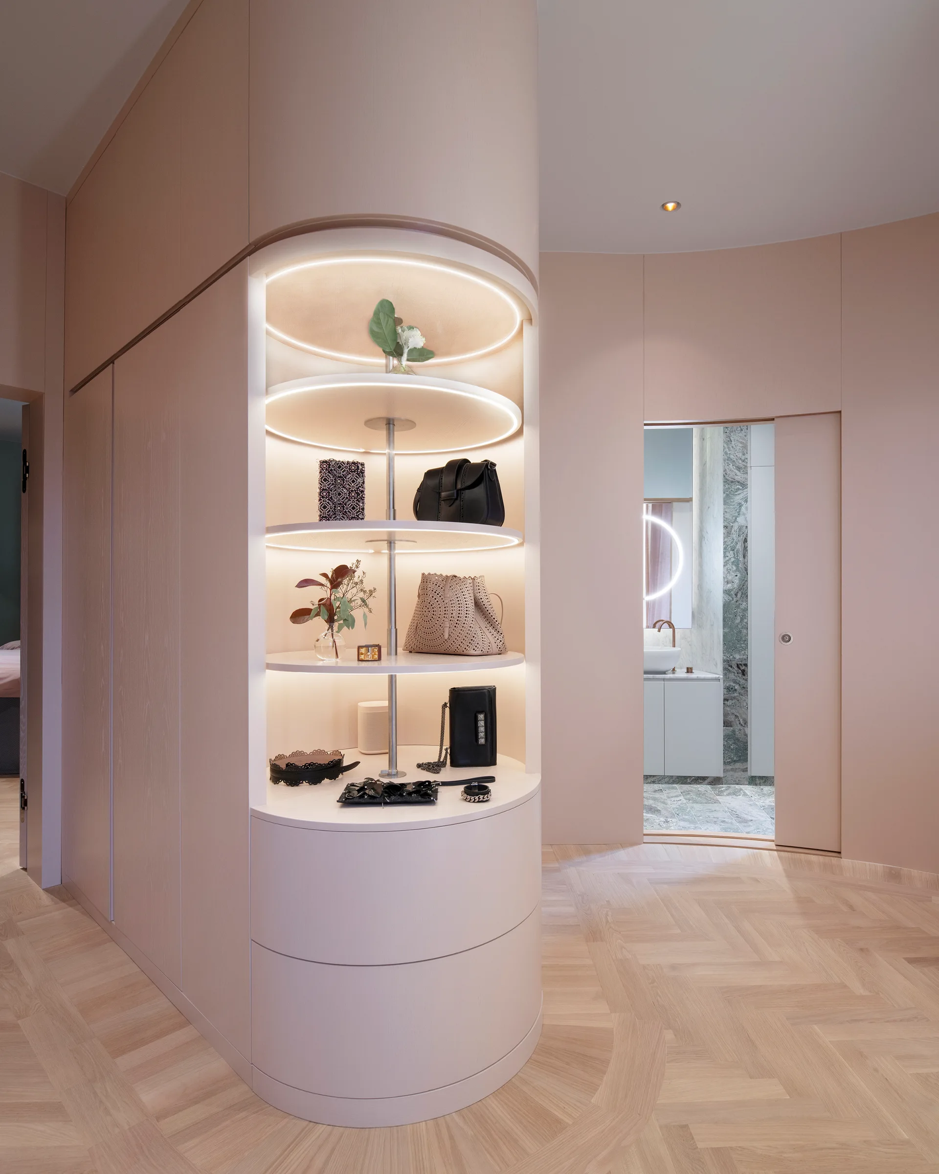 A curved pale pink wardrobe with round backlit display shelves stands on herringbone parquet wood flooring near an open sliding pocket door.