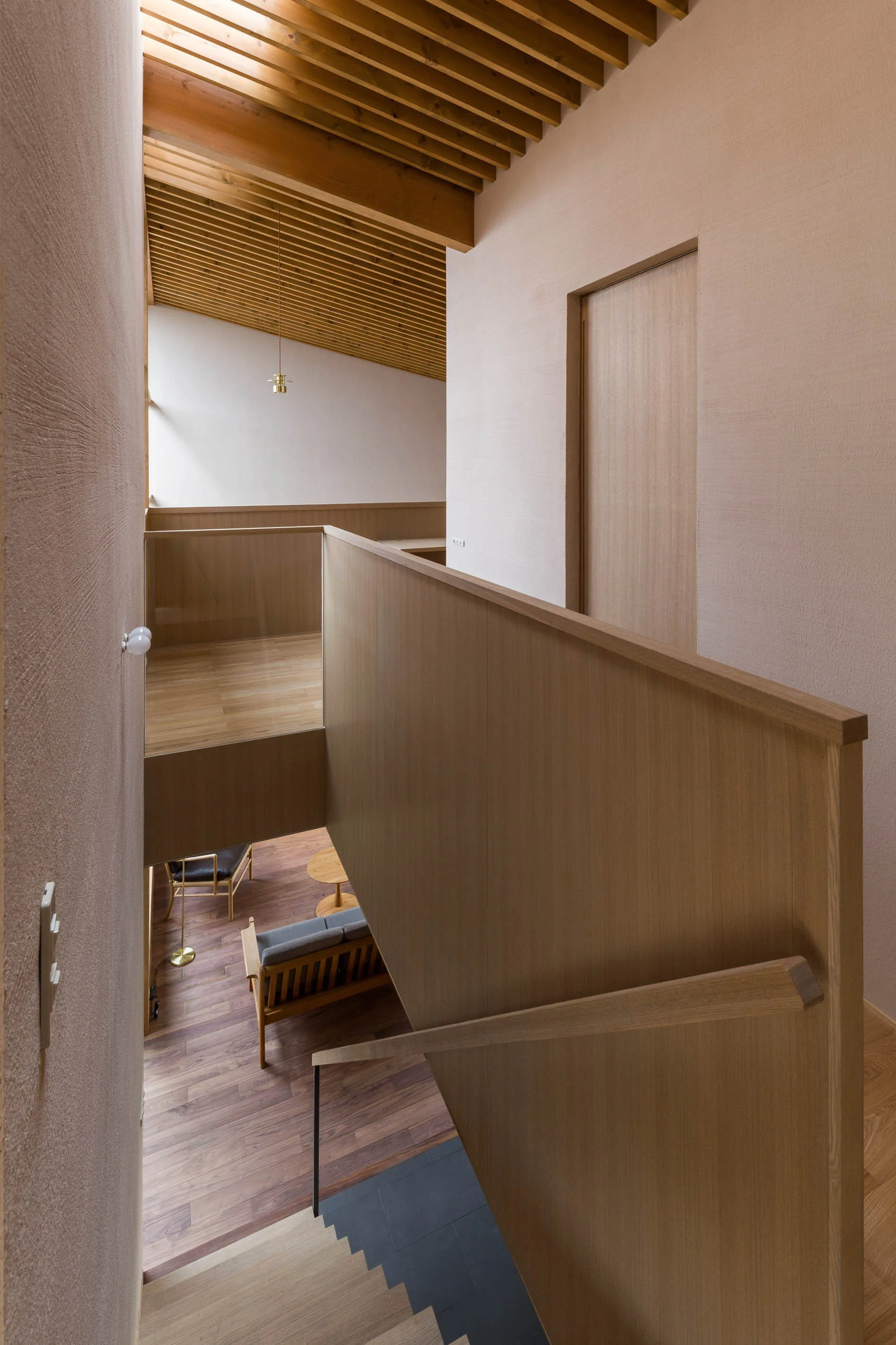 High-angle view of a wooden staircase featuring solid wood railings, textured plaster walls, and an overhead slatted timber ceiling.