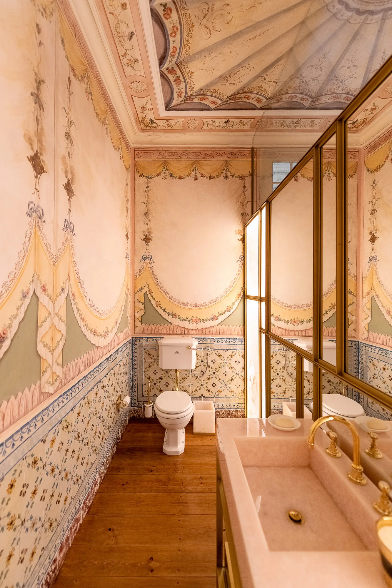 A powder room features hand-painted floral wall murals, patterned tile wainscoting, a white toilet, and a pink stone vanity with gold faucets.