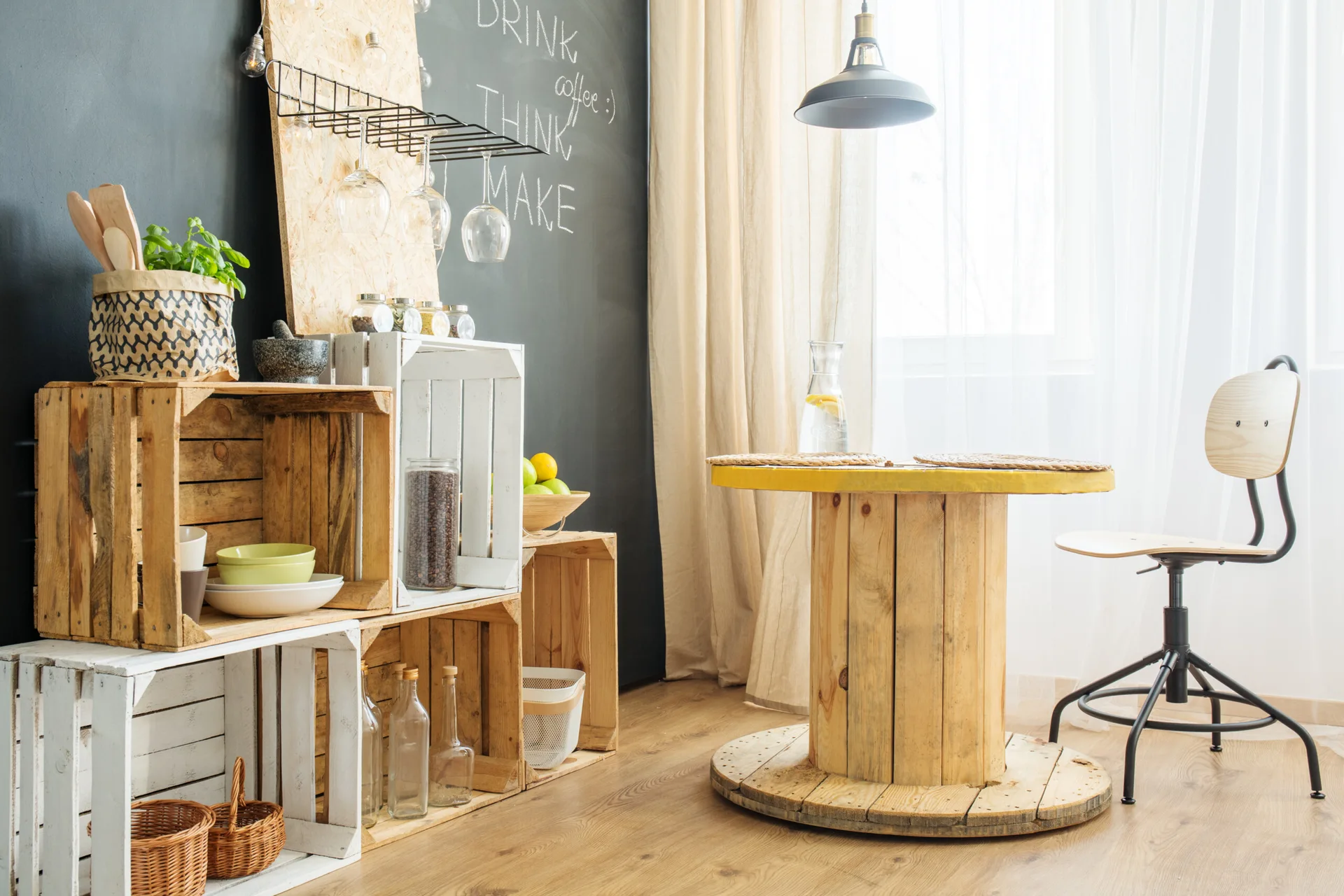 A rustic dining nook features a table made from a wooden cable reel, stacked crate shelving against a chalkboard wall, and a single chair.