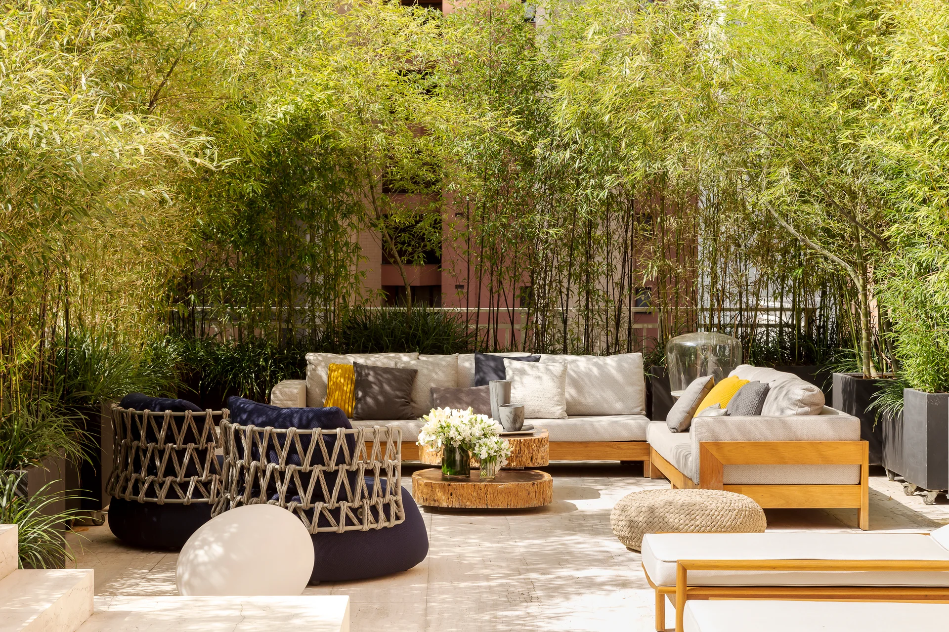 An outdoor terrace features a beige sectional sofa, navy blue woven armchairs, and wood-slab coffee tables surrounded by a tall wall of green bamboo.