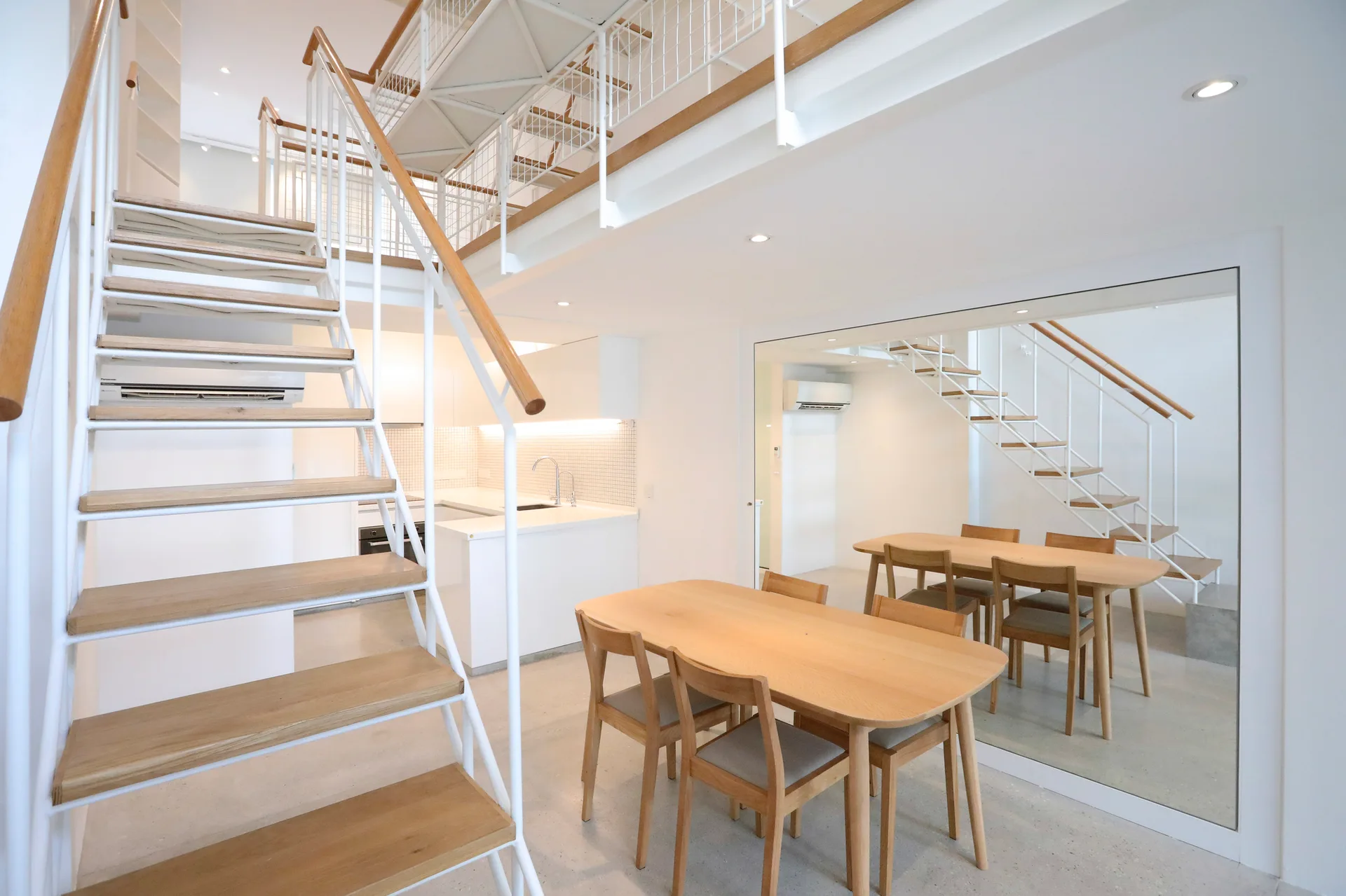 A minimalist dining area features a light wood table and chairs next to a large wall mirror and an open-riser staircase leading to a mezzanine.