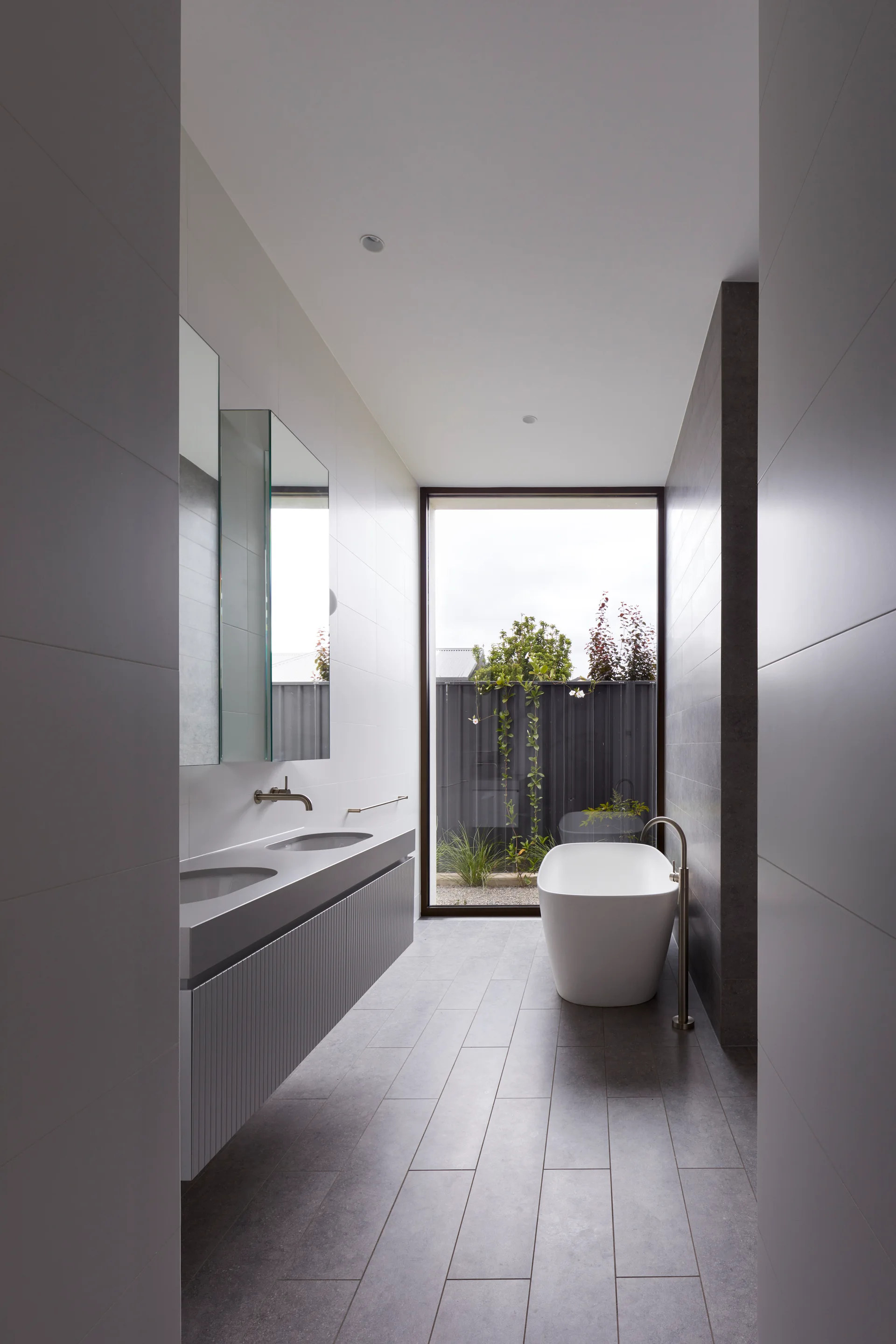 A modern minimalist bathroom featuring a gray fluted double vanity, a freestanding bathtub, and a window looking out to a courtyard.